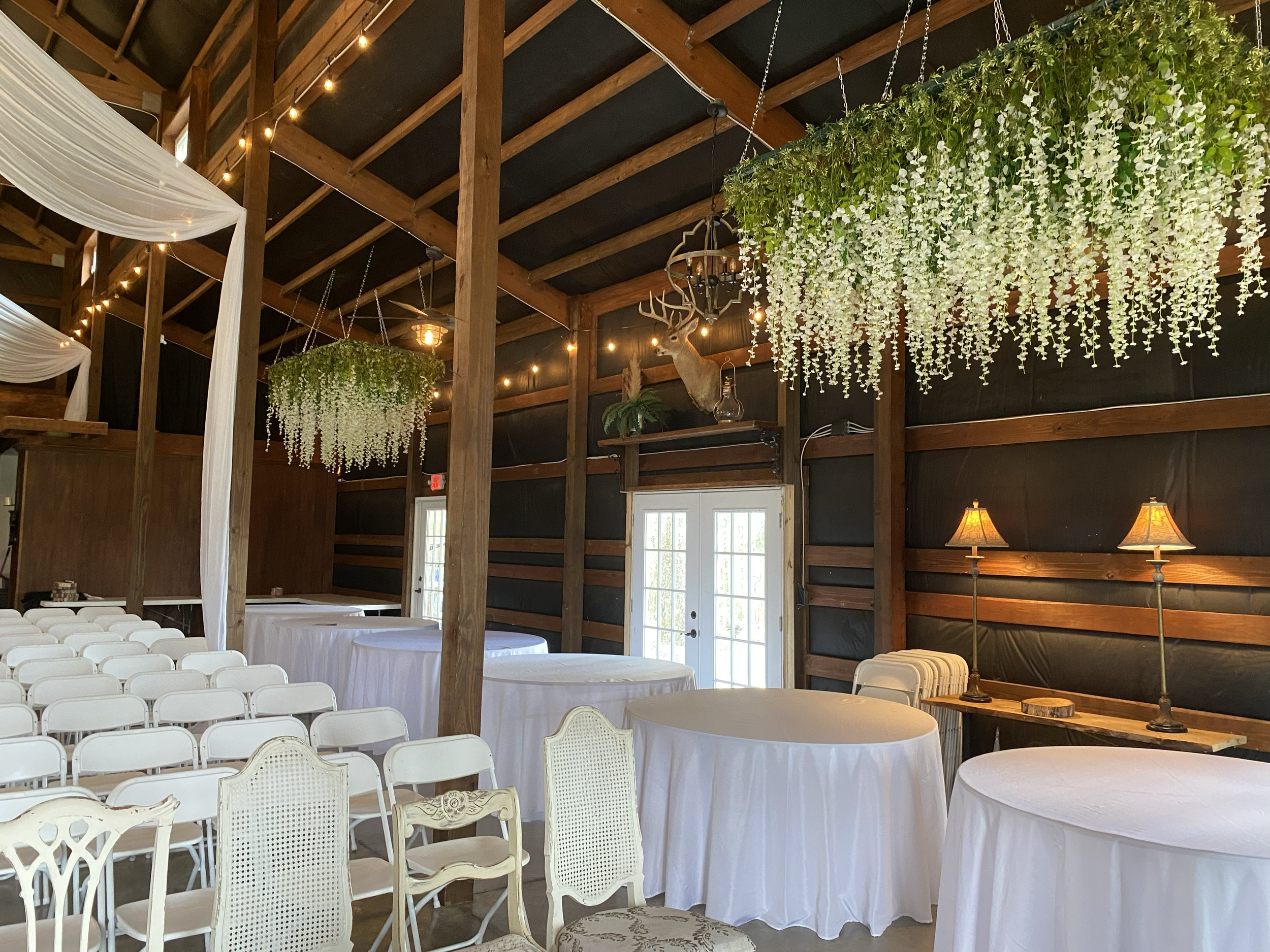 Interior of a rustic hall decorated with hanging white flowers, string lights, and a mounted deer head on the wall.