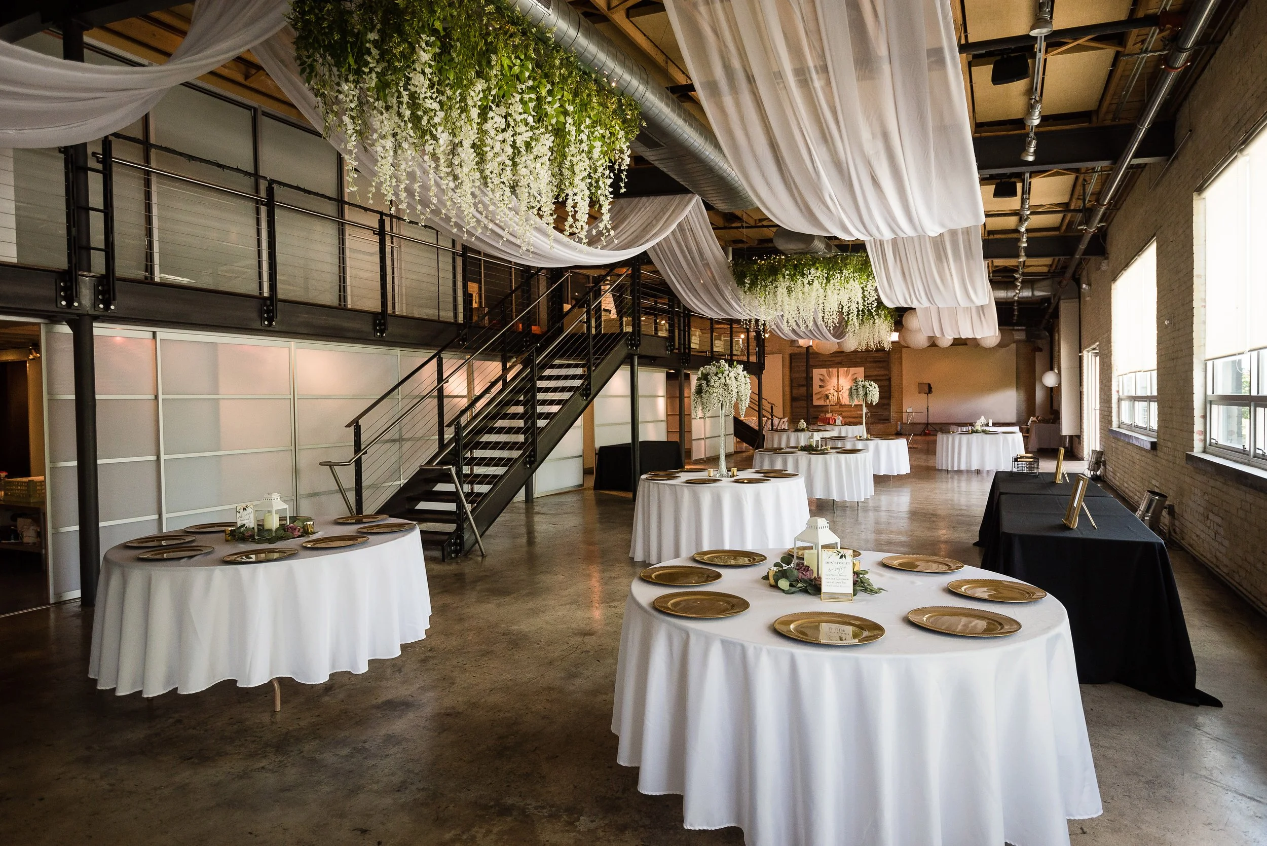 Interior of a decorated event space with round tables covered in white tablecloths, gold plates, and centerpieces, and ceiling drapes with hanging white flowers and greenery.