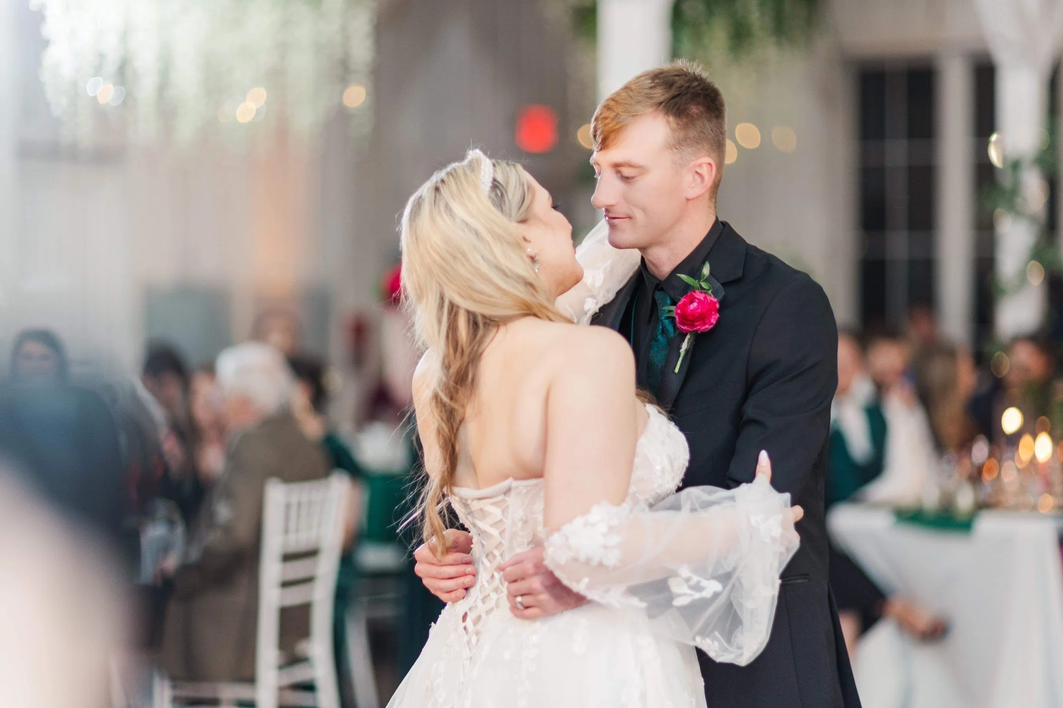 A bride and groom dancing at their wedding reception, with guests seated at tables in the background.