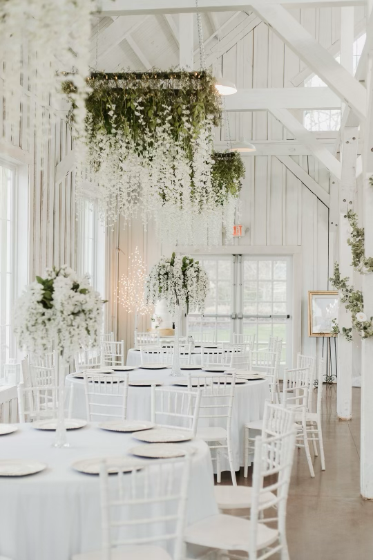 A bright, airy wedding or event reception with white tables, chairs, and floral centerpieces, decorated with hanging greenery and flowers in a white rustic wooden barn-style venue.