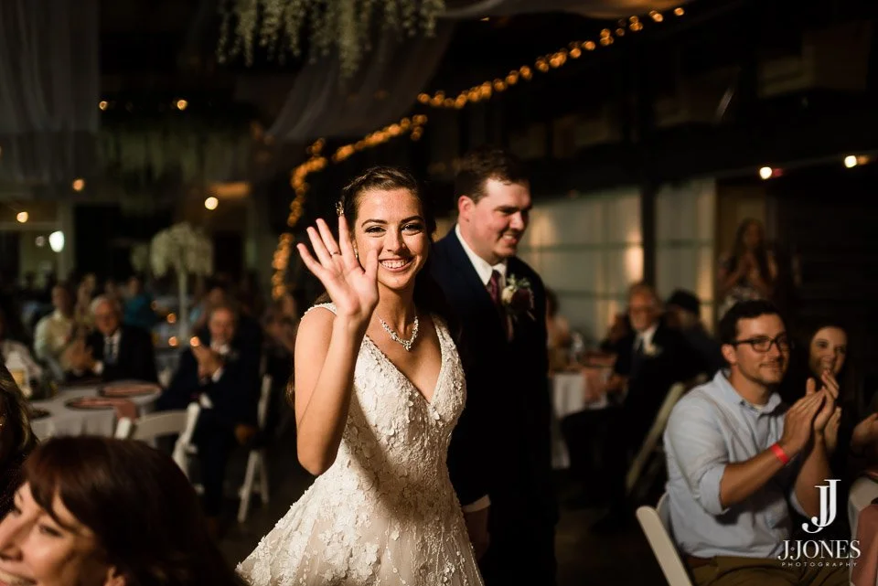 A bride with a wedding dress waving at the camera in a wedding reception, with guests clapping in the background.