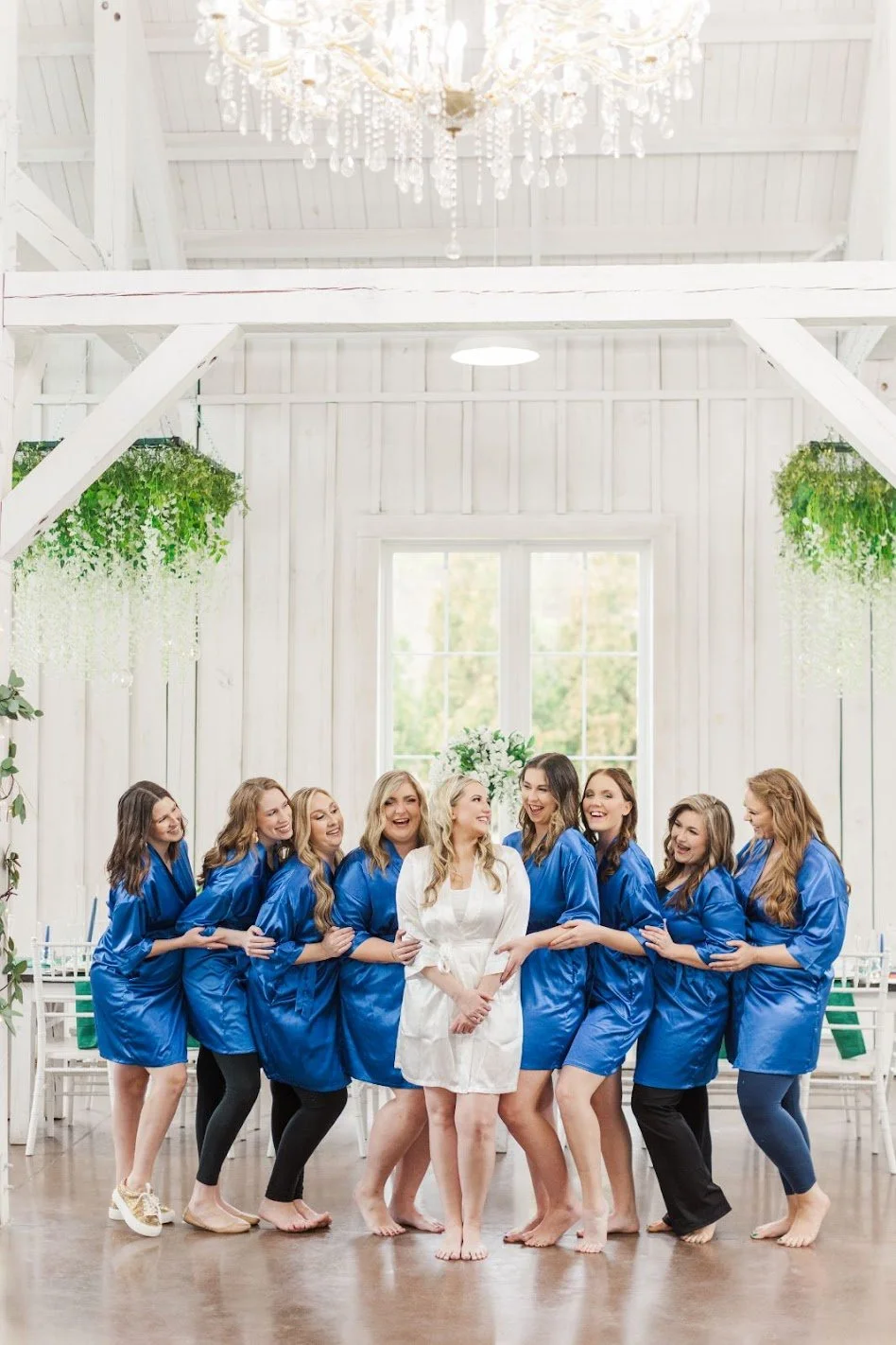 A group of women in blue robes, with one woman in a white robe, laughing and smiling together in a bright, white room with hanging greenery and a large chandelier.