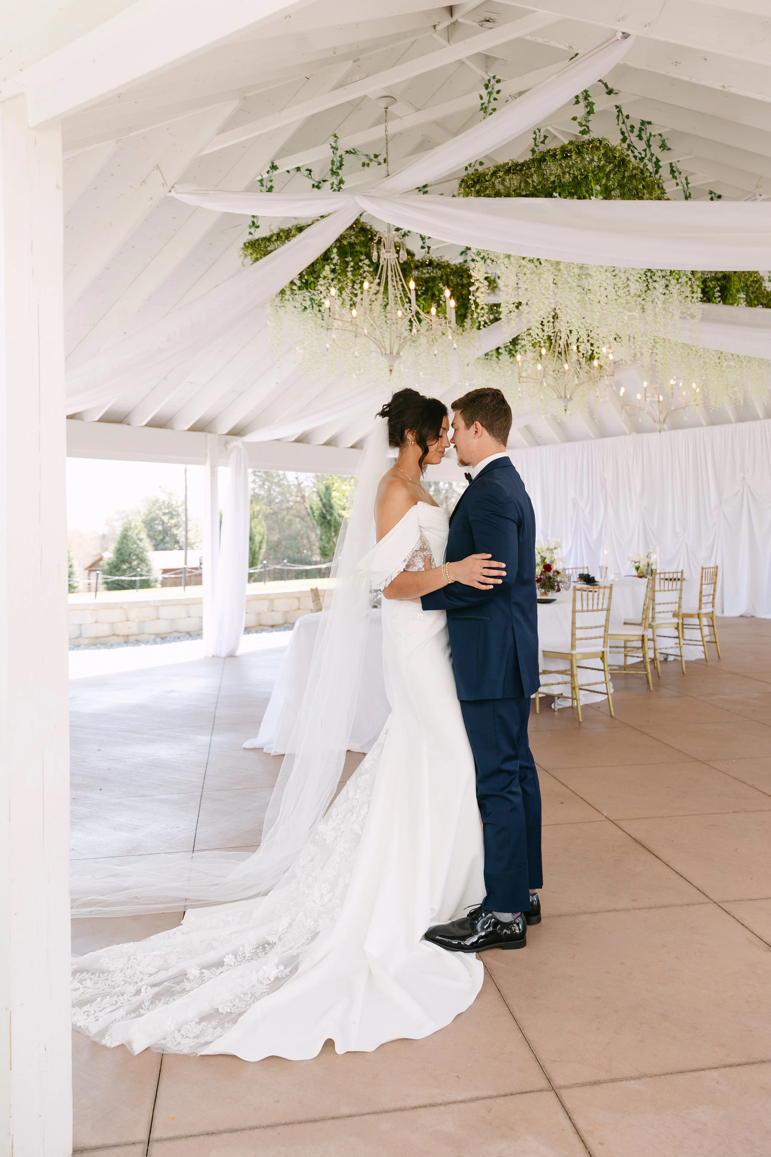 A bride and groom are holding each other closely during their wedding ceremony in a decorated outdoor pavilion. The bride wears a white wedding dress with lace details and a long train, and the groom wears a navy blue suit with black shoes. The ceili