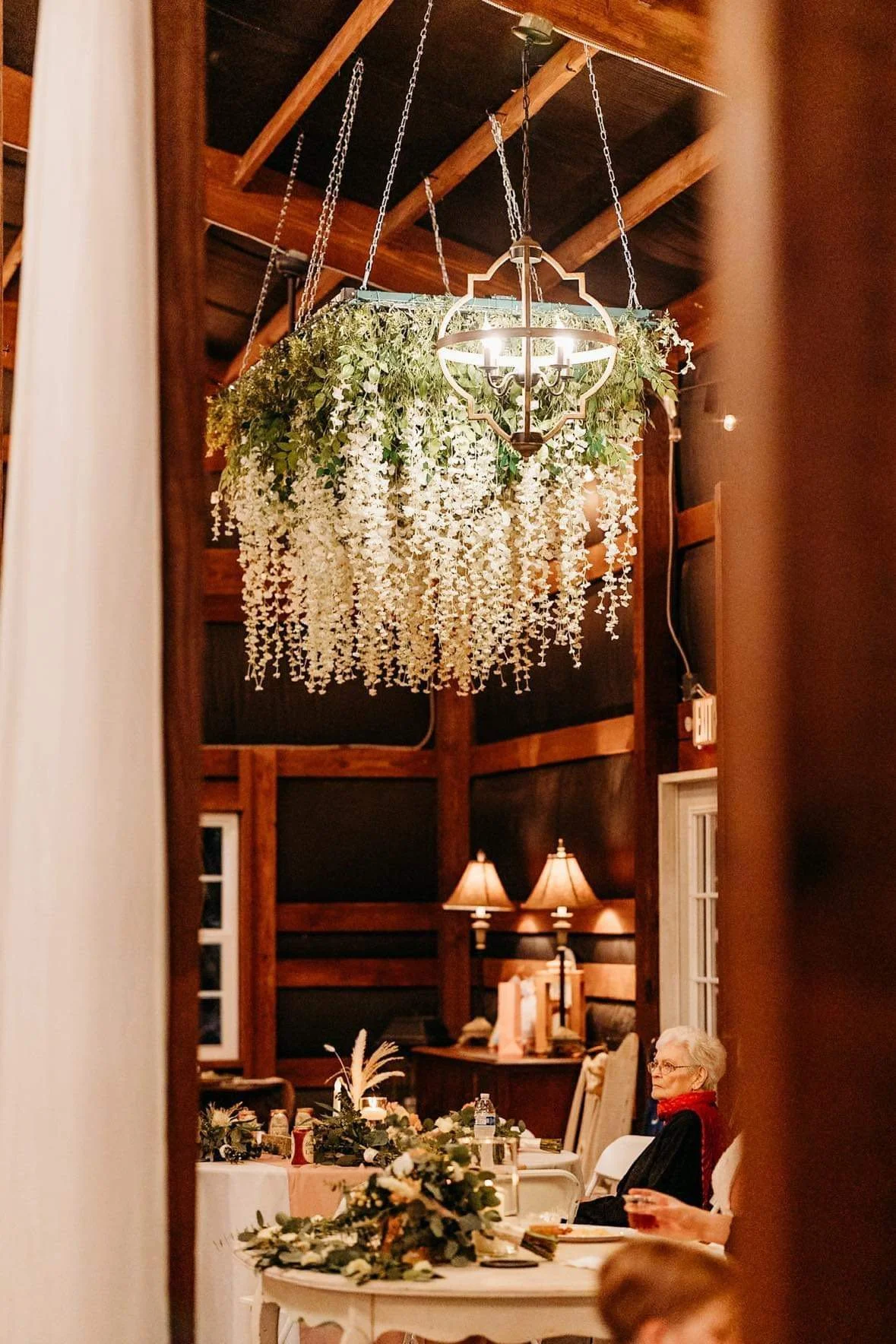 A woman seated at a decorated dining table during a festive gathering, with a large floral chandelier and warm lighting in the background.