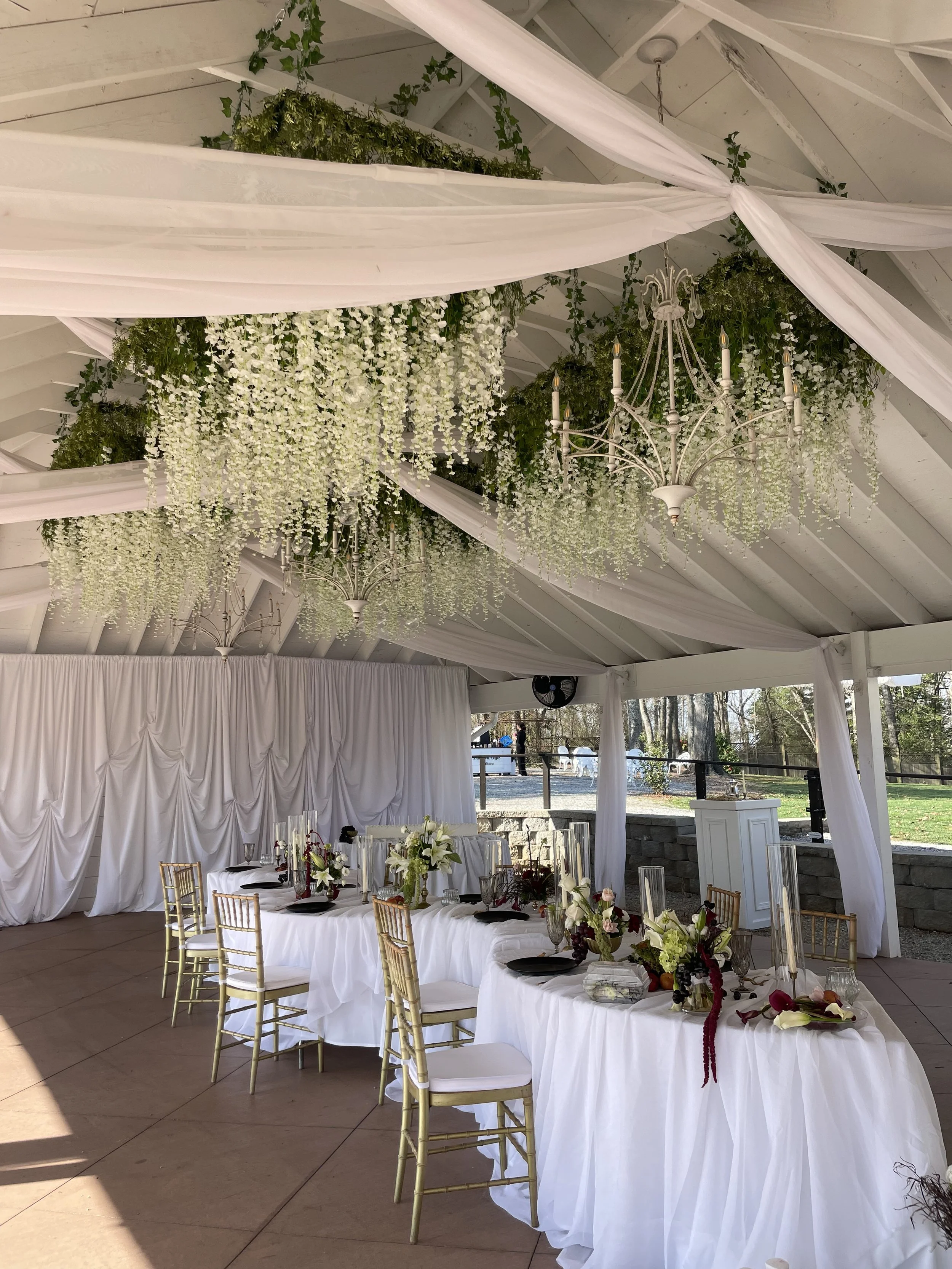 Elegant outdoor event space decorated with white drapes, floral arrangements, and chandeliers, featuring a long table with gold chairs and a white curtain backdrop.