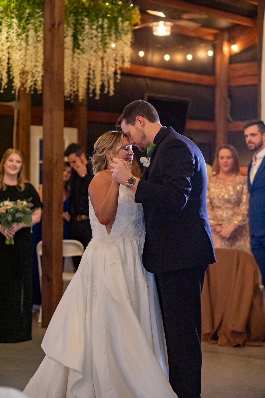 A bride and groom share a dance at their wedding reception, surrounded by friends and family, in a warmly lit venue with wooden accents and floral decorations.