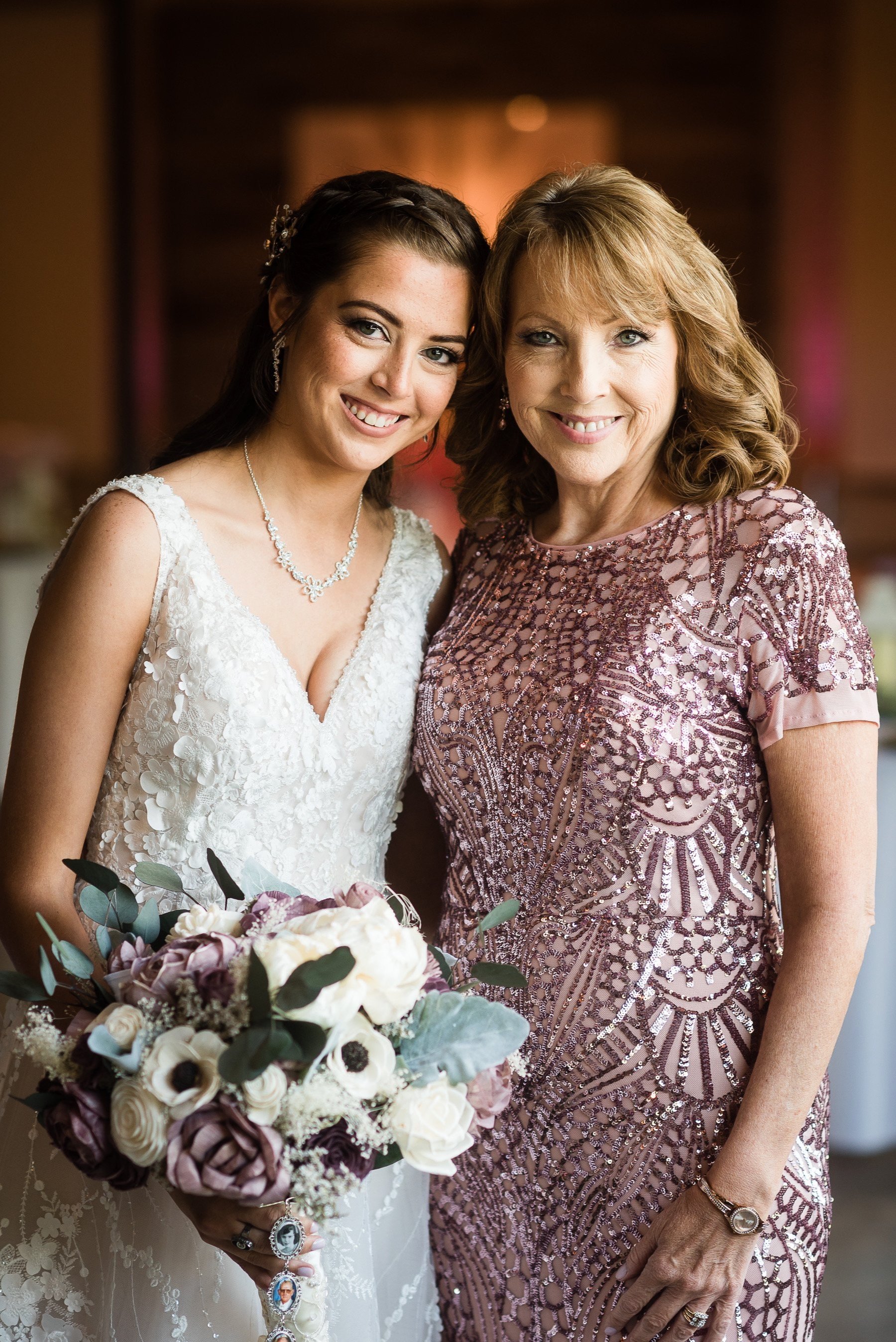 A young woman in a white lace wedding dress holding a bouquet of flowers, standing next to an older woman in a pink patterned dress, both smiling at the camera.