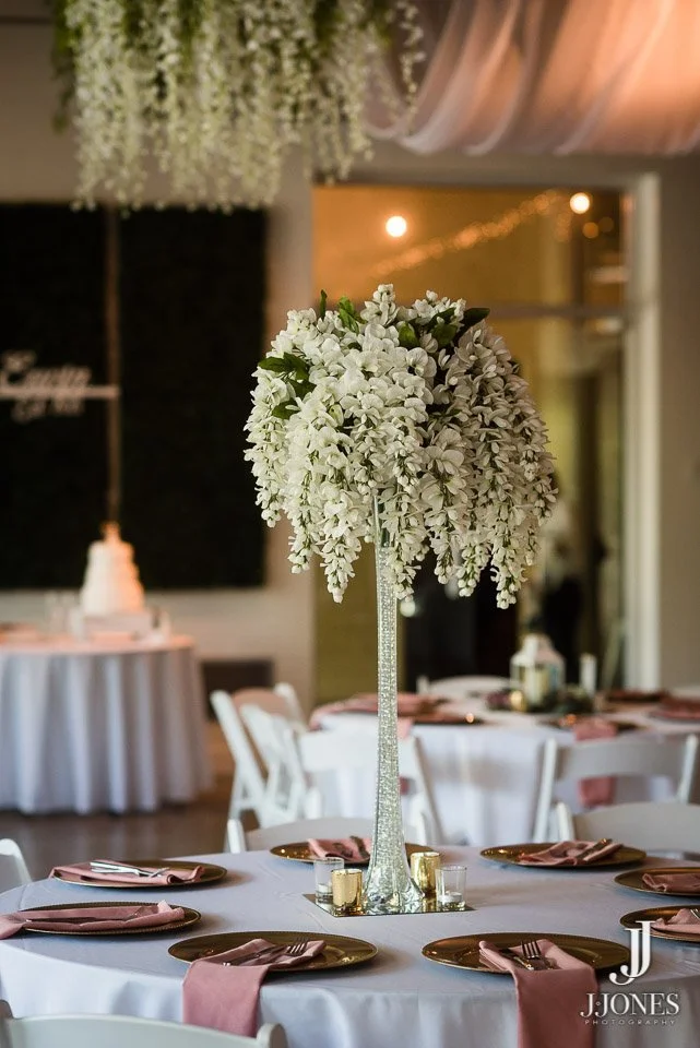 Elegant event table with a tall silver floral centerpiece of white cascading flowers, with pink napkins and gold plates arranged around the table, in a decorated venue with soft lighting.