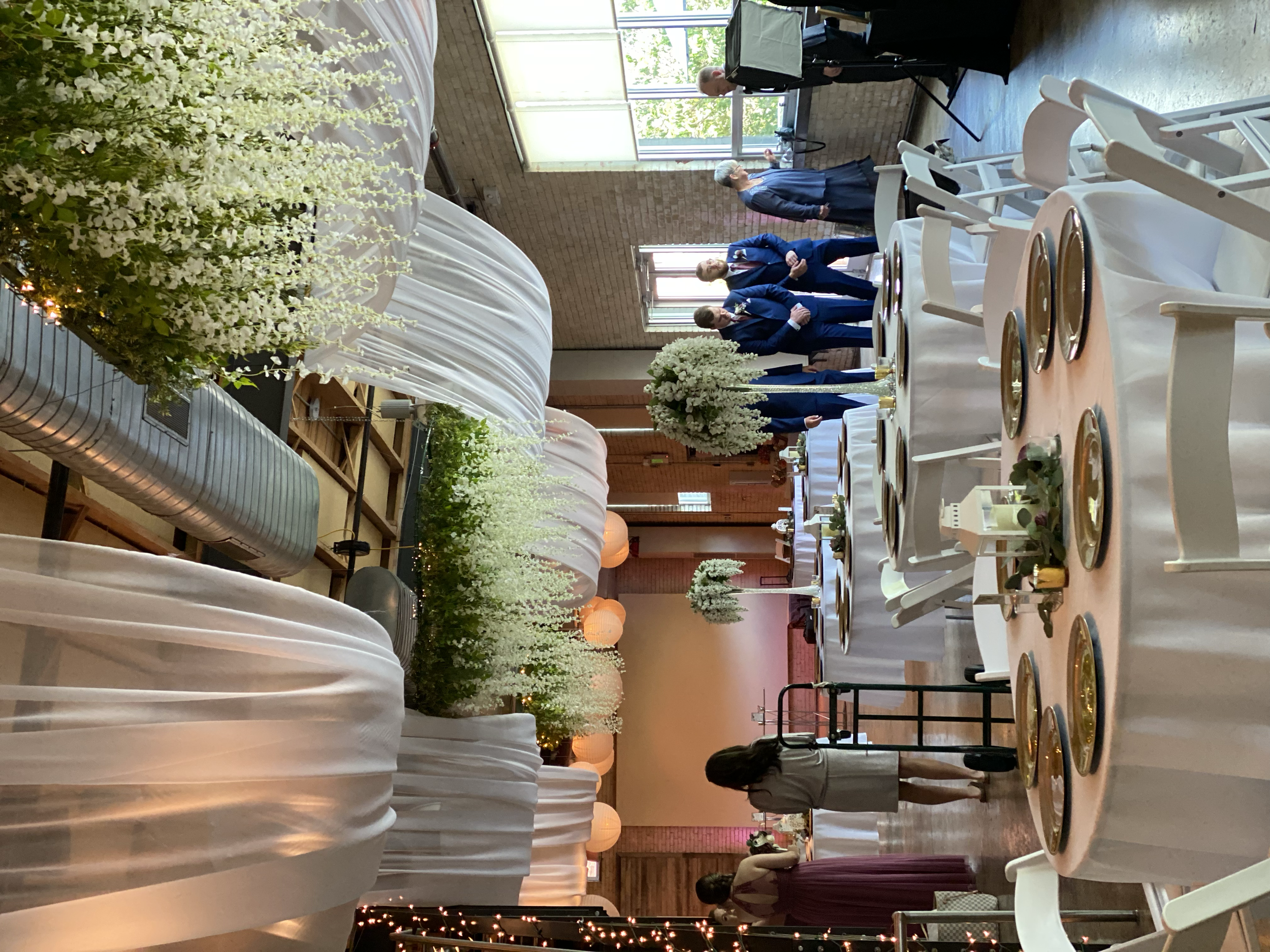 Inside a decorated event space, a decorated cake and floral arrangements are visible, with guests in formal attire mingling and talking. There are tables set with plates and cutlery, and large windows letting in natural light.