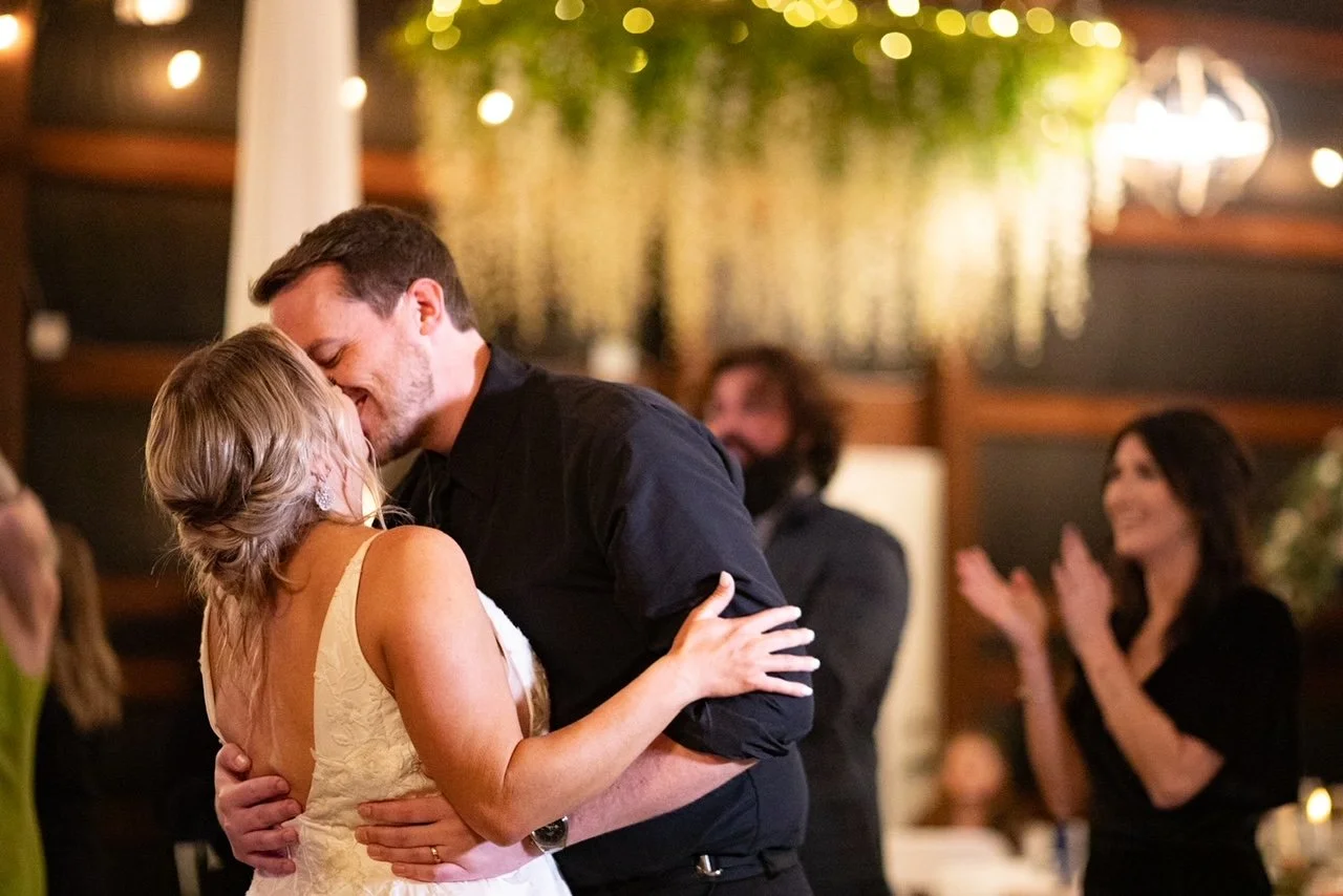 A couple dancing closely at a wedding reception, smiling and sharing an intimate moment, with guests clapping in the background.