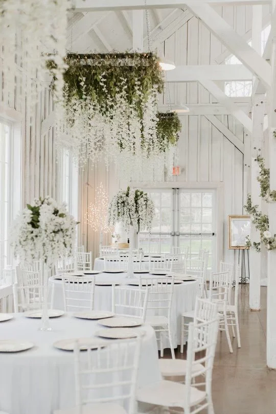Elegant white wedding reception setup with round tables, white chairs, floral centerpieces, hanging greenery, and bright natural lighting in a rustic barn-style venue.