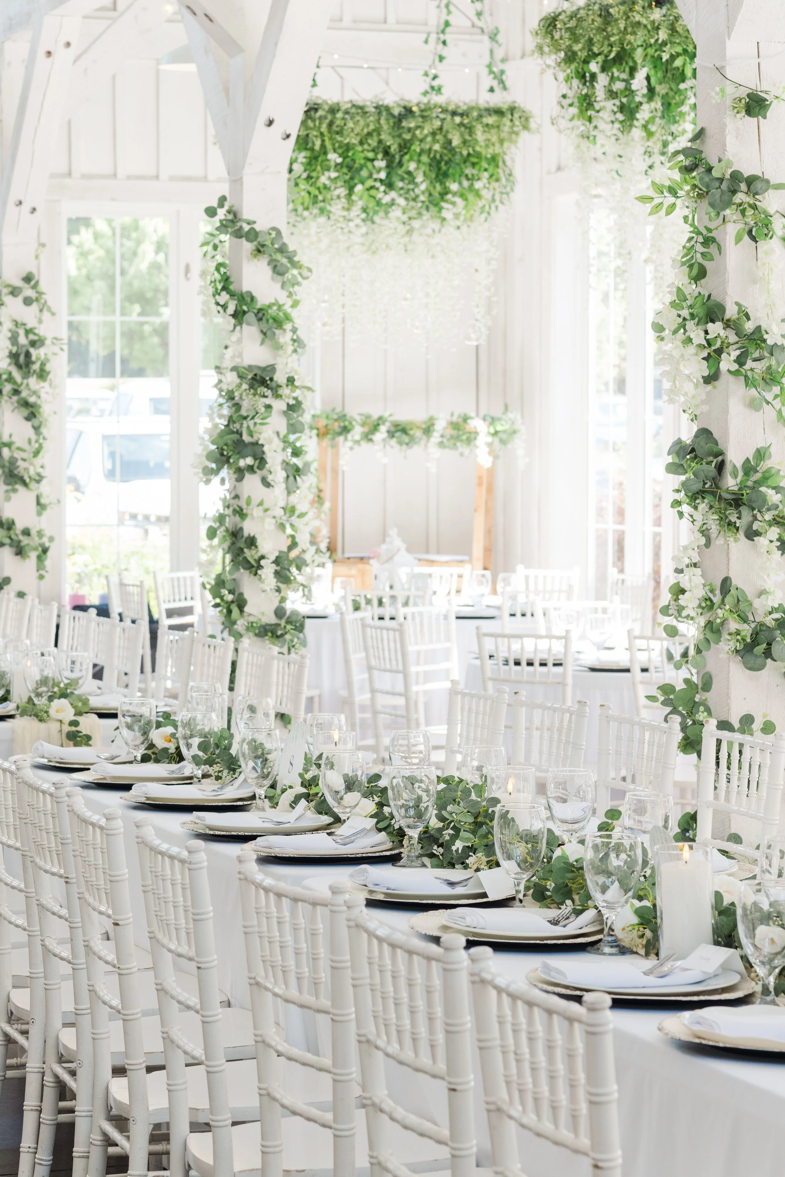 Elegant wedding reception setup with white chairs, round tables, glassware, and floral arrangements, inside a bright venue with large windows and hanging greenery.