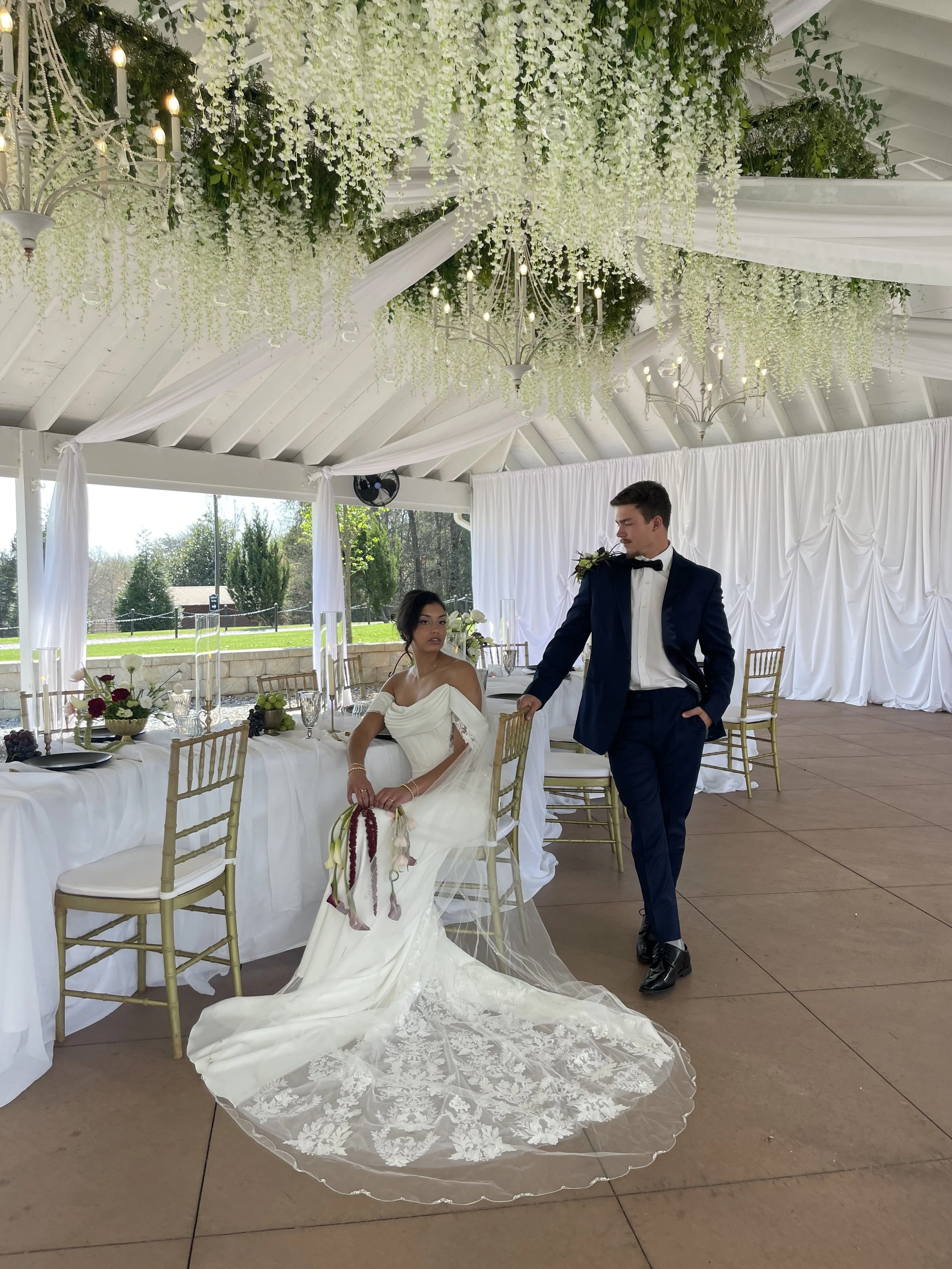 A bride and groom at a wedding reception indoors with white drapery and hanging floral arrangements.