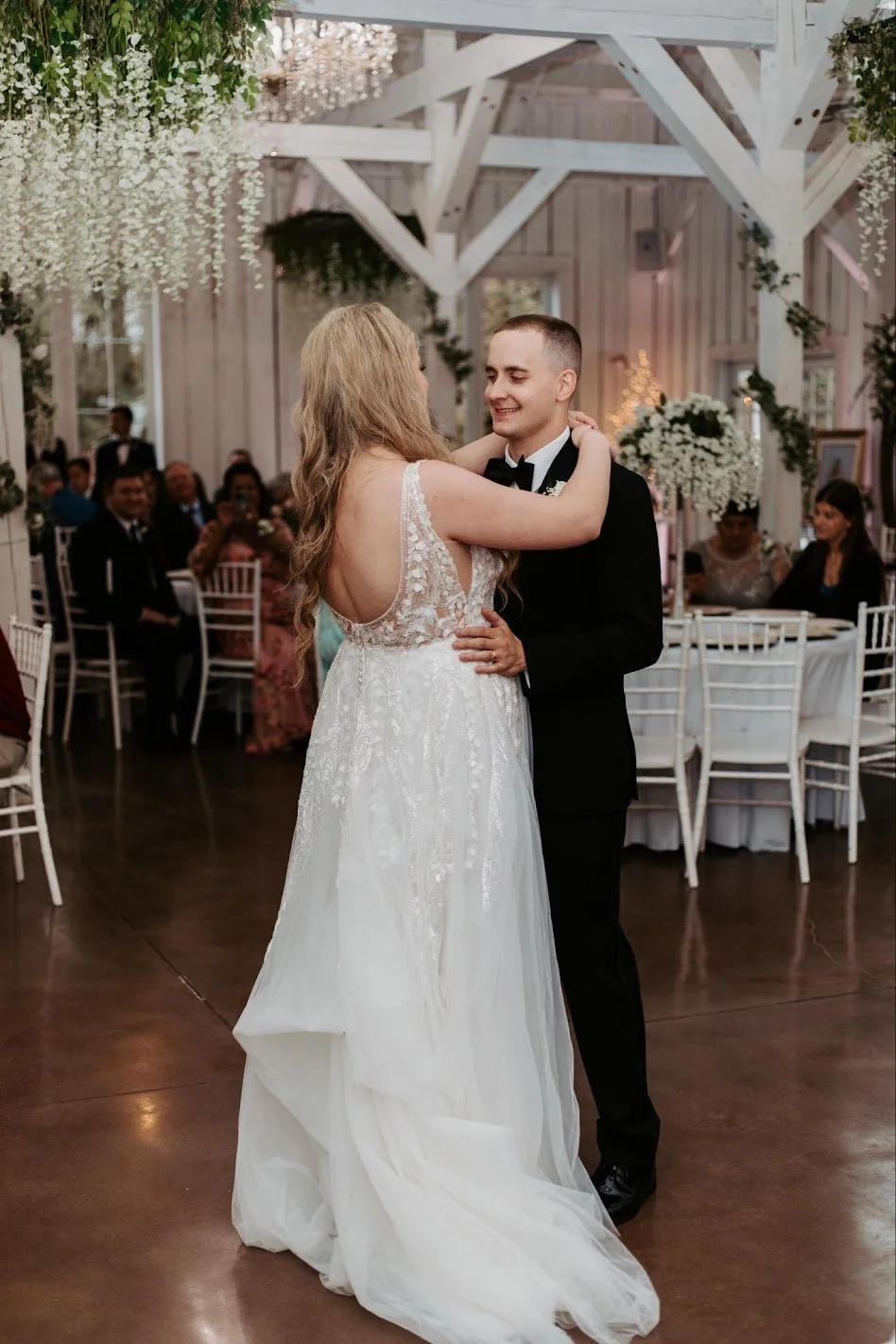 A bride and groom dancing at their wedding reception in a decorated venue with hanging flowers and seated guests watching.
