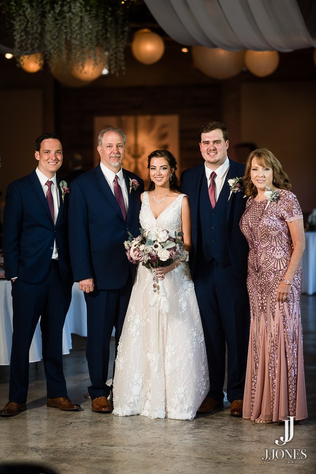 Group of five people standing together at a wedding reception, with the bride in a white gown holding a bouquet, and the groom in a suit, flanked by family members in formal attire, inside a decorated venue with hanging lights.