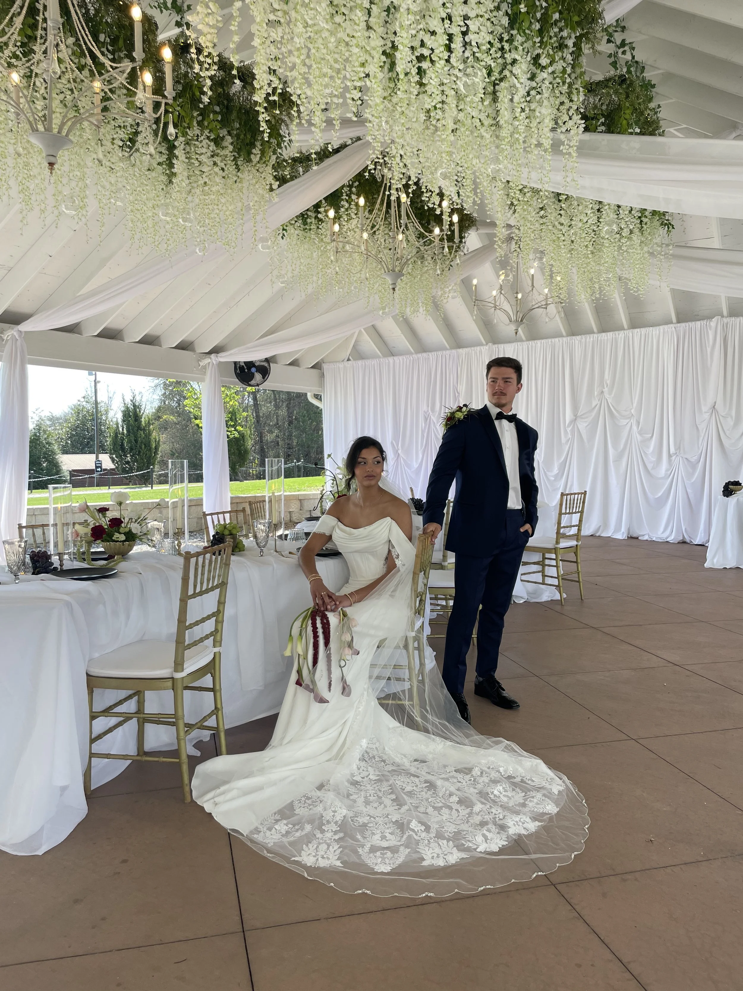 A bride and groom at a wedding reception, indoors with white drapery and floral decorations hanging from the ceiling, and a decorated table with fruit and flowers.