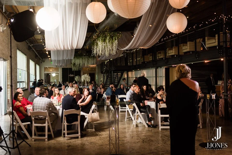 Guests seated at round tables with white tablecloths in a decorated event space with hanging paper lanterns and draped fabric, attending a formal gathering or celebration.