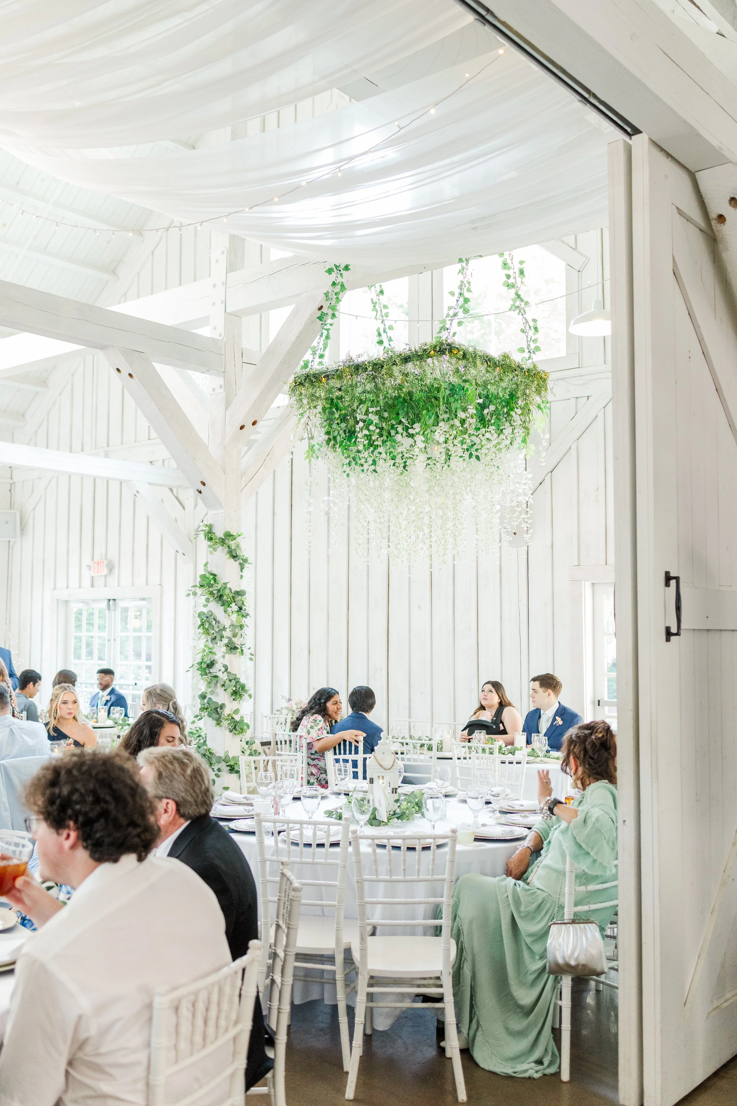 A wedding reception inside a bright, white rustic barn with high ceilings, wooden beams, and hanging greenery and flowers. Guests sit at round tables with white chairs, engaging in conversation and dining.