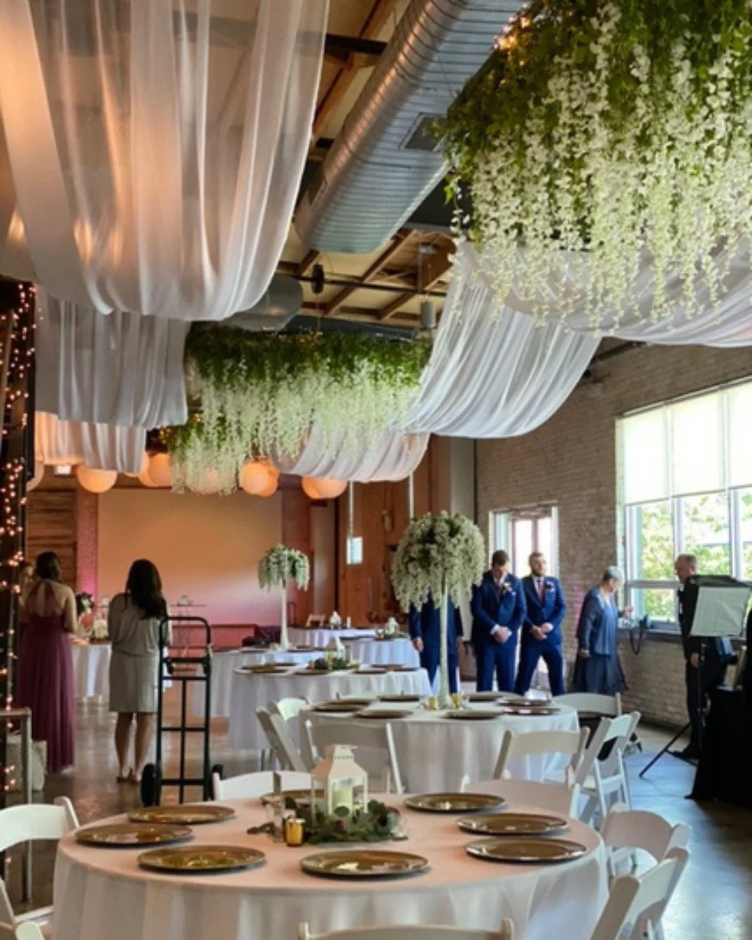 An indoor wedding reception decorated with white drapes, hanging greenery, and floral centerpieces; round tables set with plates and candles, with a stage and people preparing in the background.