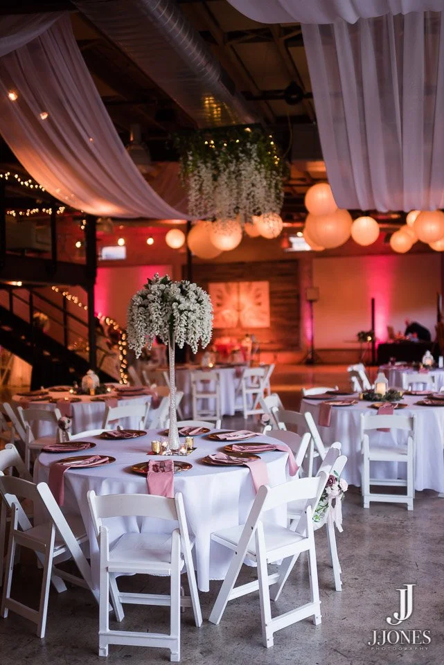Wedding reception with round tables covered in white tablecloths, decorated with pink napkins, small candles, and floral centerpieces. White chairs surround each table. The ceiling is adorned with white drapery and hanging round paper lanterns, with 