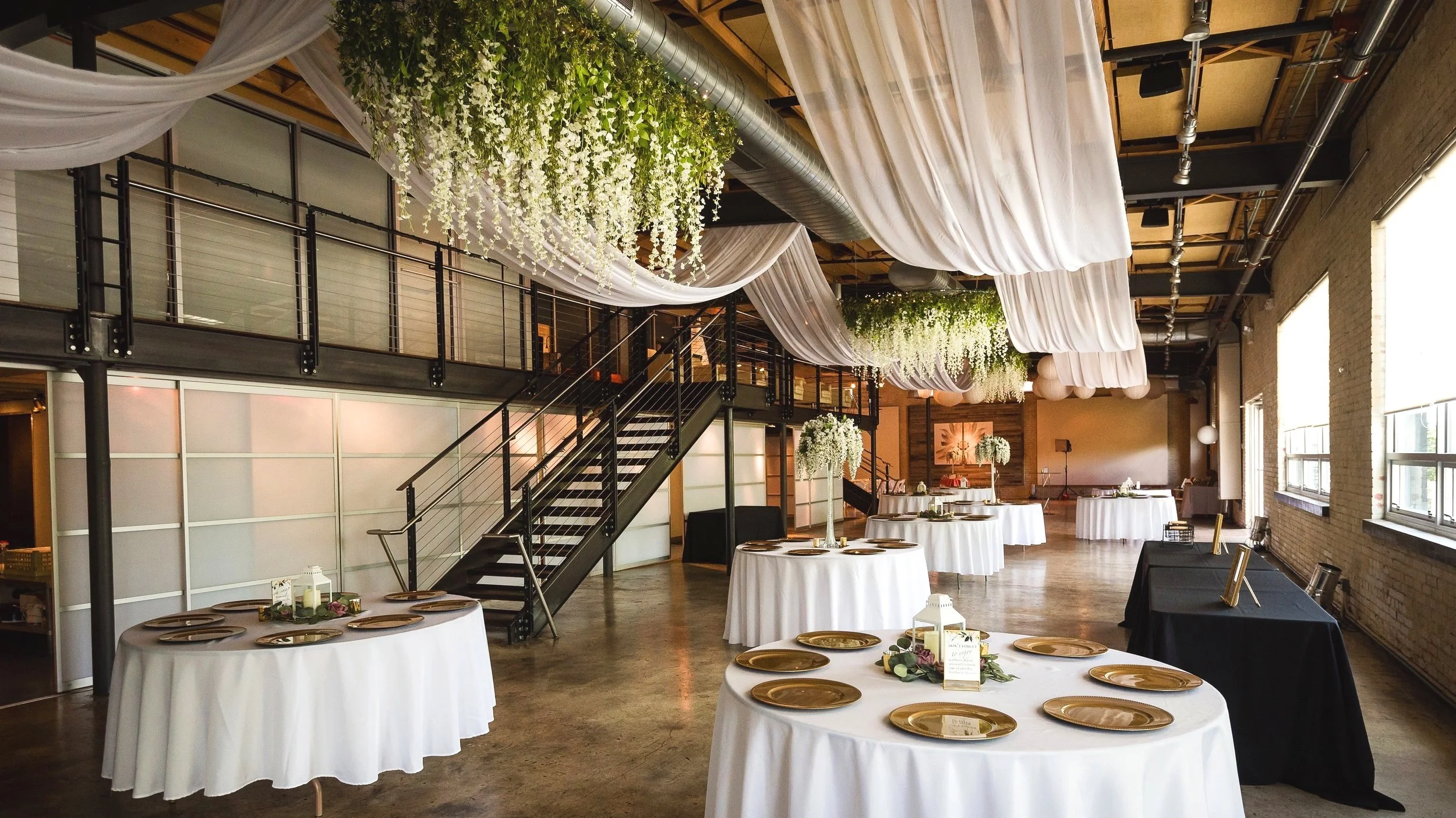 Wedding reception setup in an industrial-style venue with round tables covered in white tablecloths, gold chargers, and floral centerpieces. White drapery and hanging greenery decorate the ceiling, with a staircase and windows in the background.