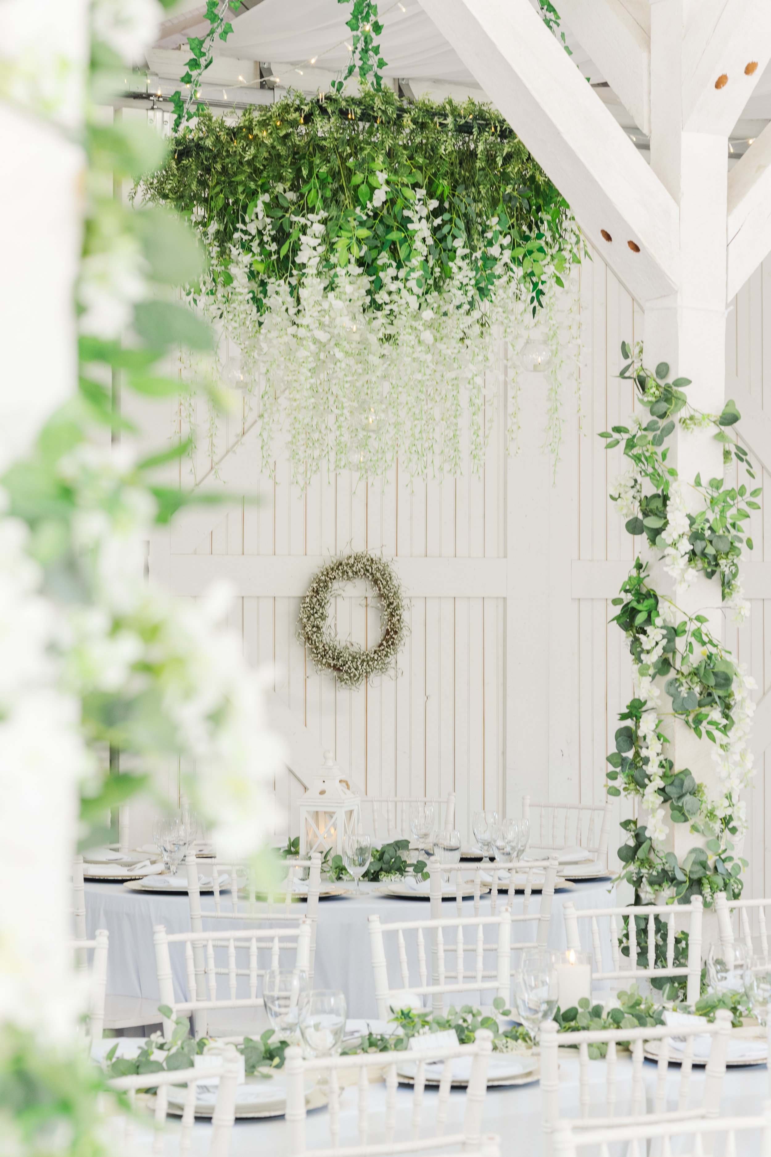 A decorated event space with hanging greenery, white flowers, and string lights above a round table set with white chairs, glassware, and a lantern in the center.