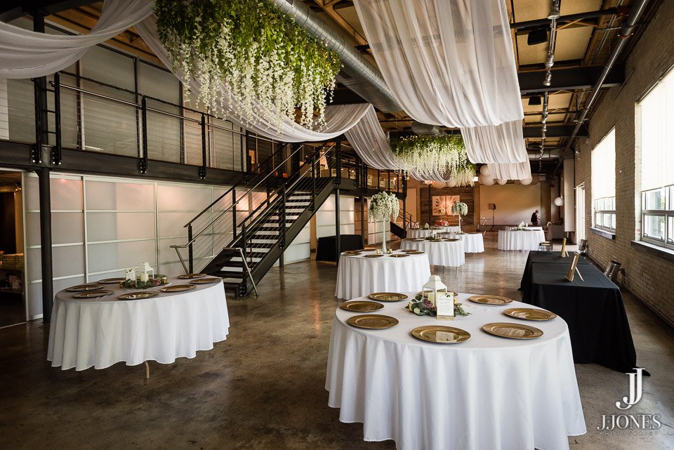 Event space decorated with draped white fabric and hanging greenery, featuring round tables with white tablecloths and gold plates for dining.