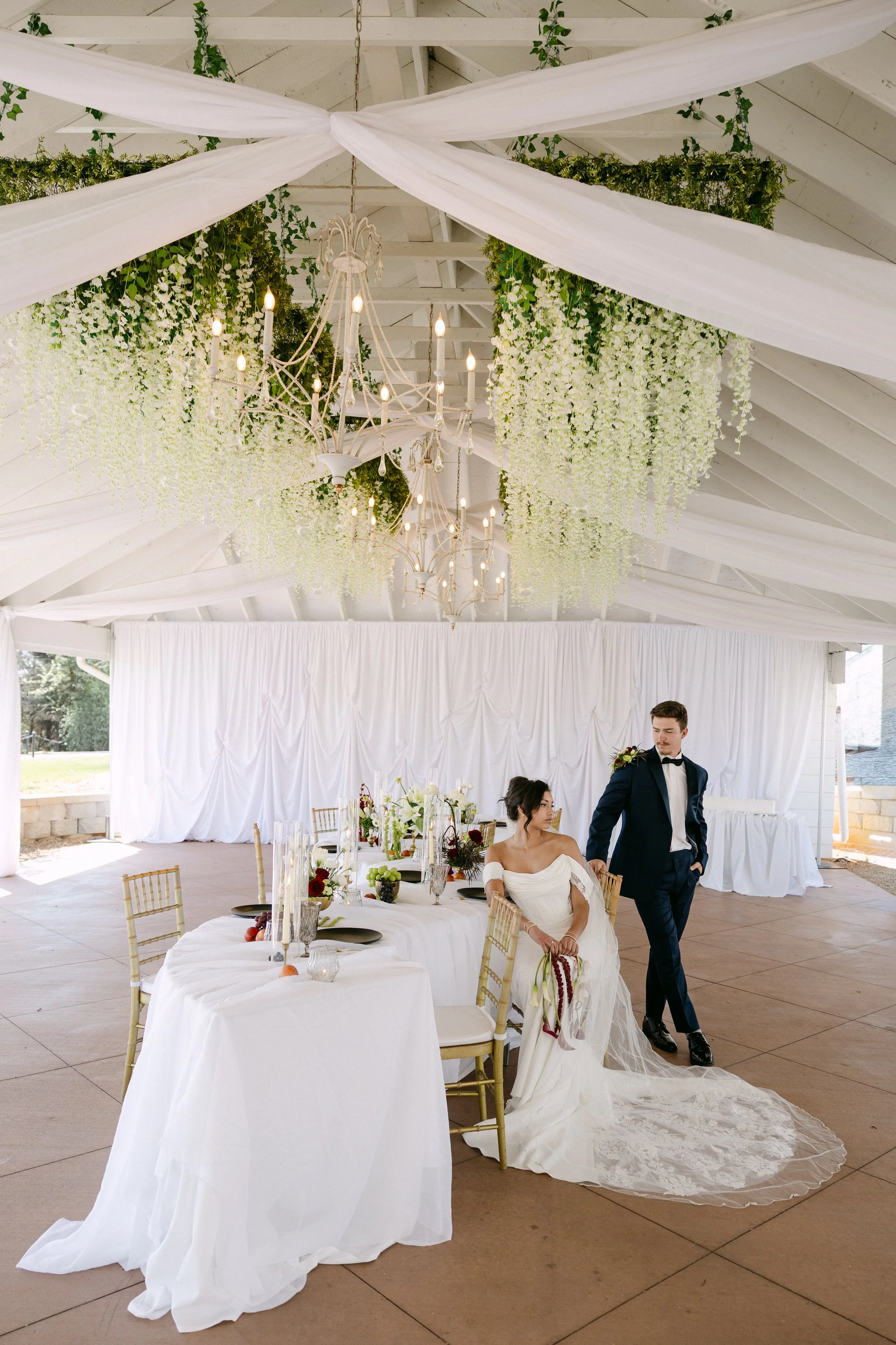 Wedding reception with a long table set with white tablecloths, fruit, and floral arrangements. A bride in a white gown is sitting, and a groom in a dark tuxedo is standing beside her. The ceiling is decorated with white drapes, greenery, and hanging