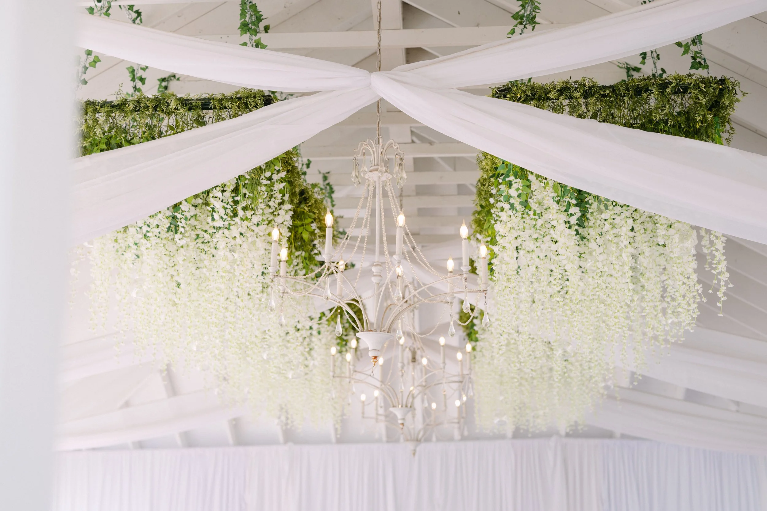 White ceiling decorated with flowing white drapes, hanging greenery, and a white chandelier with lit candles.