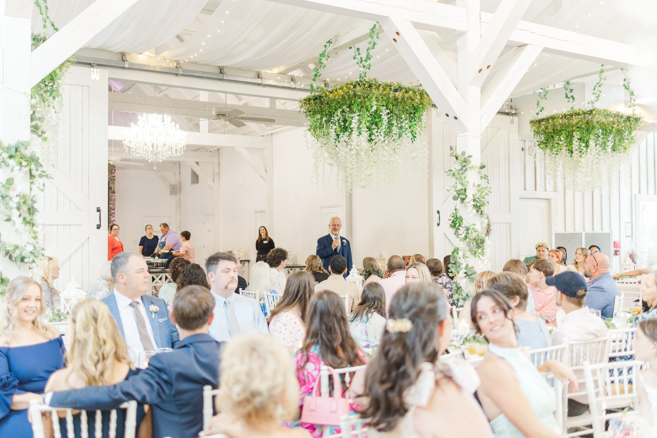 A wedding reception with guests seated at tables, a man in a suit giving a speech, decorative greenery and hanging floral arrangements, a chandelier, and white drapery on the ceiling in a bright, rustic venue.