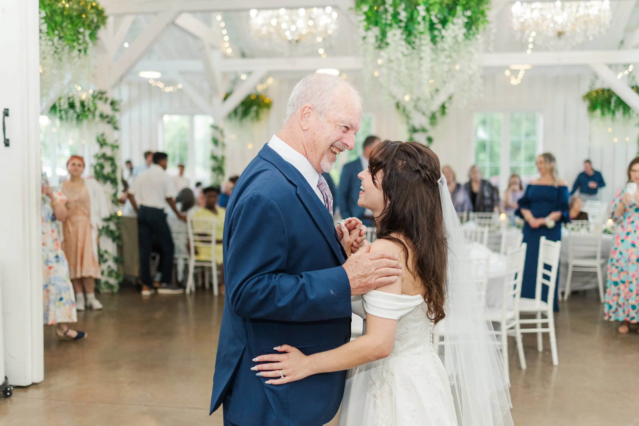 An elderly man in a blue suit and a young woman in a white wedding dress are dancing and smiling at each other at a wedding reception inside a brightly decorated venue with greenery and hanging lights.