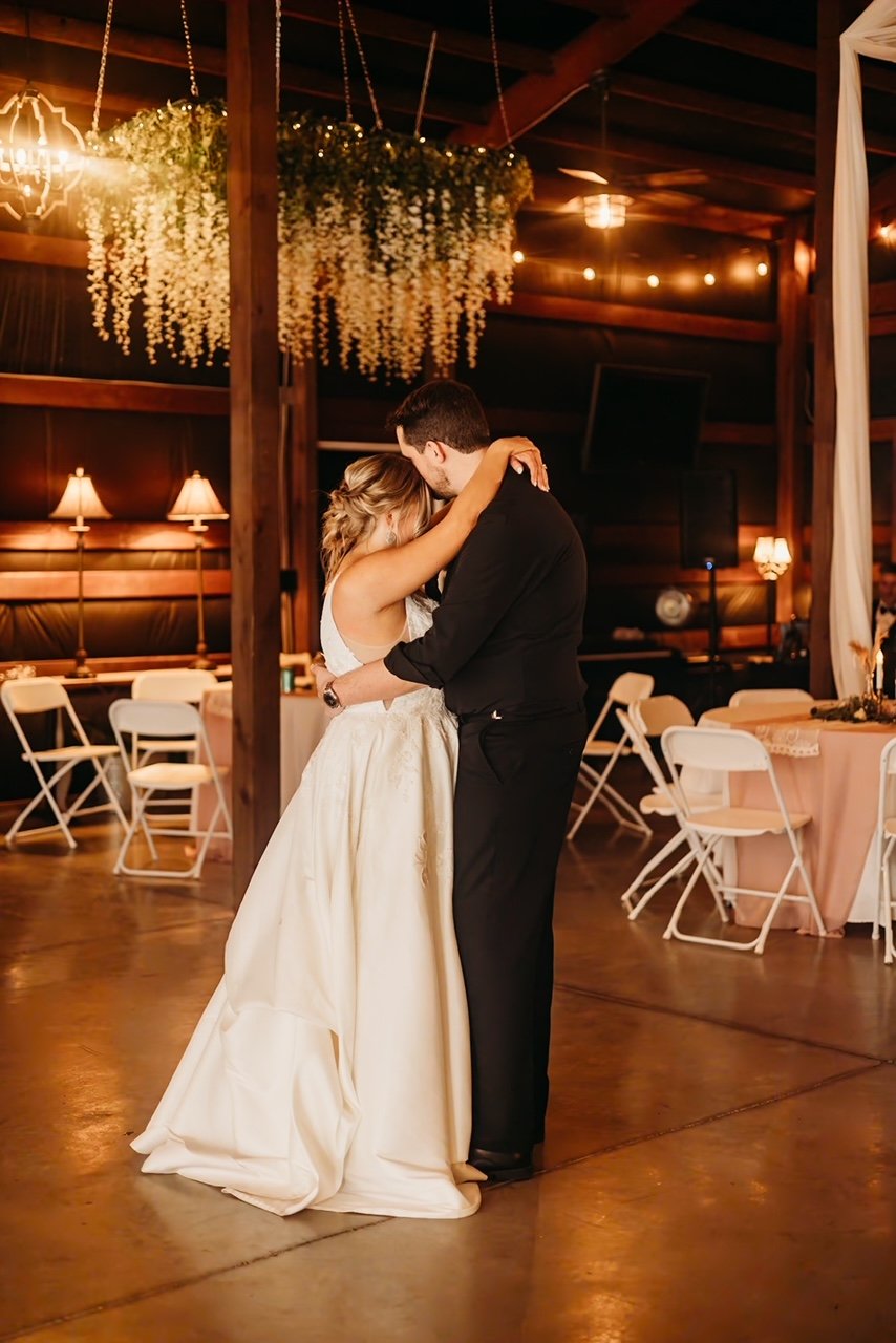 A couple shares a slow dance at a wedding reception in a warmly lit venue with wooden interior and floral decorations.