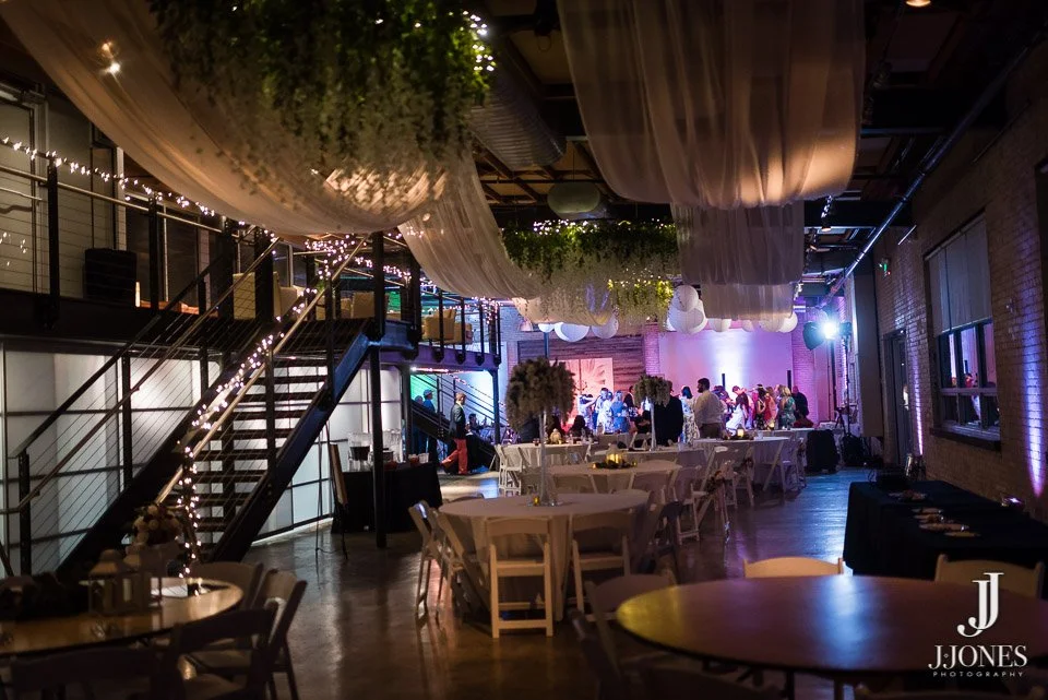 Indoor event space decorated with hanging drapes and fairy lights, with a dance floor and a group of people dancing in the background, tables and chairs set up for dining, and ambient uplighting on the walls.