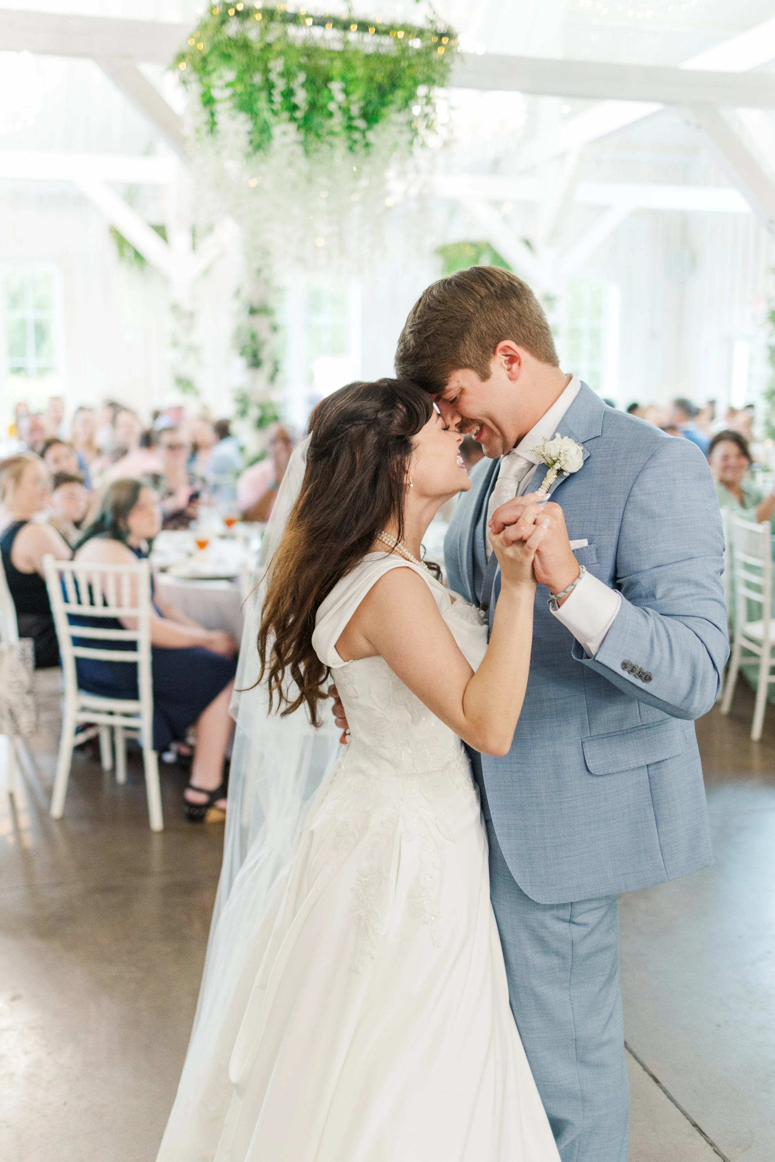 A bride and groom share a dance at their wedding reception. The bride is wearing a white wedding gown, and the groom is dressed in a light blue suit. They are smiling and holding hands, with the bride's veil visible. Guests are seated at tables in the background, and the venue is decorated with hanging greenery and fairy lights.