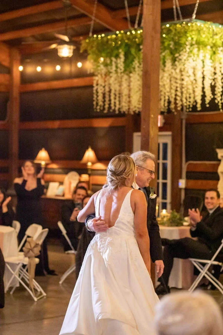 A bride and groom dancing at their wedding reception, with guests clapping and taking pictures in a warmly lit, rustic wooden venue decorated with hanging greenery and string lights.