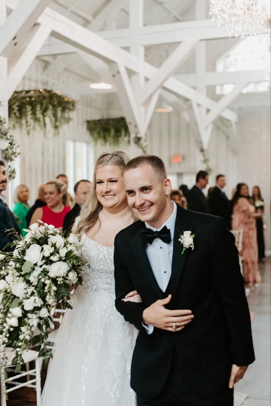 A bride and groom smiling at their wedding reception with guests in the background.