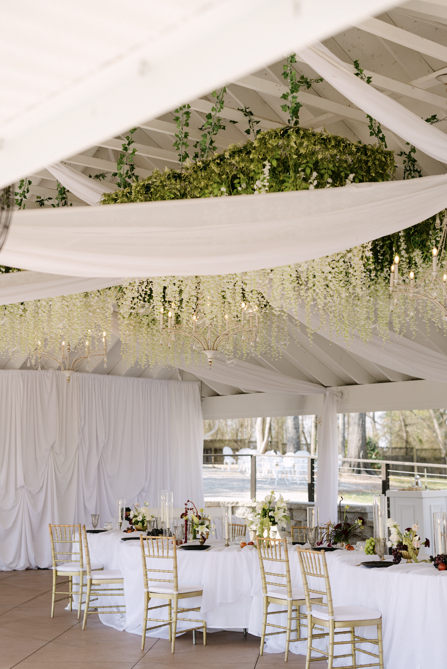 Elegant event space with white draped fabric, floral ceiling decorations, and a long dining table with white tablecloths and gold chairs.