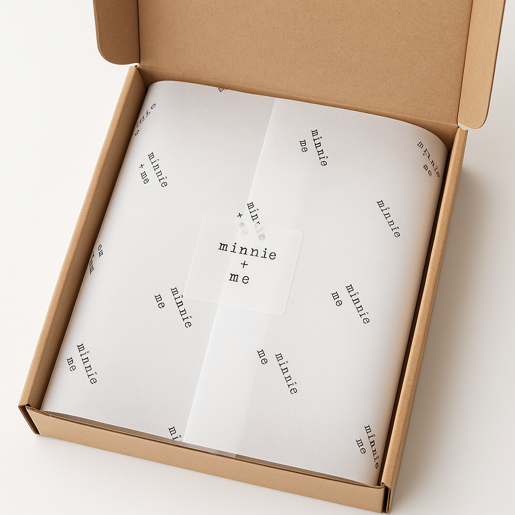 Open kraft gift box with white tissue paper printed with “minnie + me” in black typewriter font, sealed with a matching branded sticker.