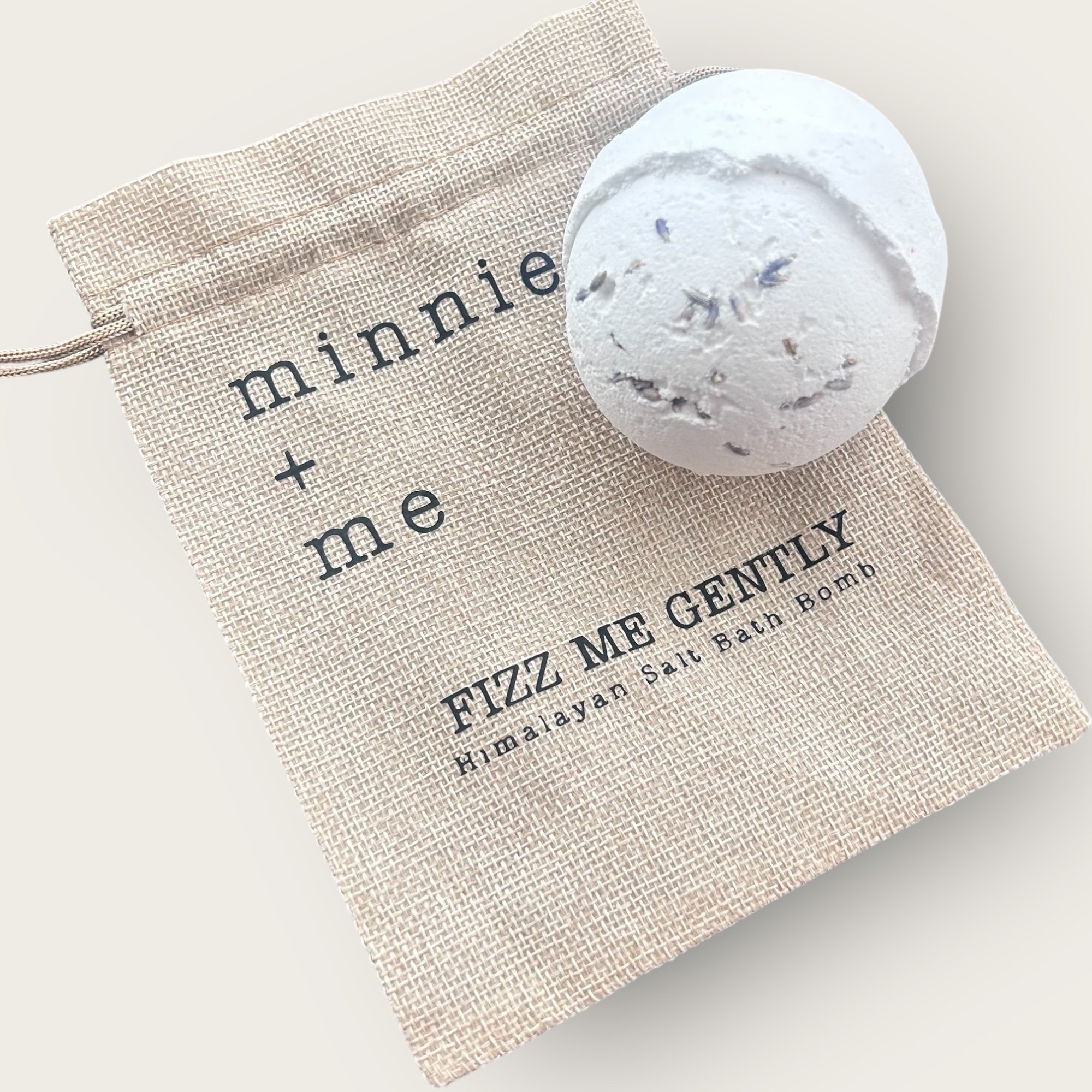 A white bath bomb with dried lavender on top, placed on a beige cloth bag labeled 'minnie + me FIZZ ME GENTLY Himalayan Salt Bath Bomb'.