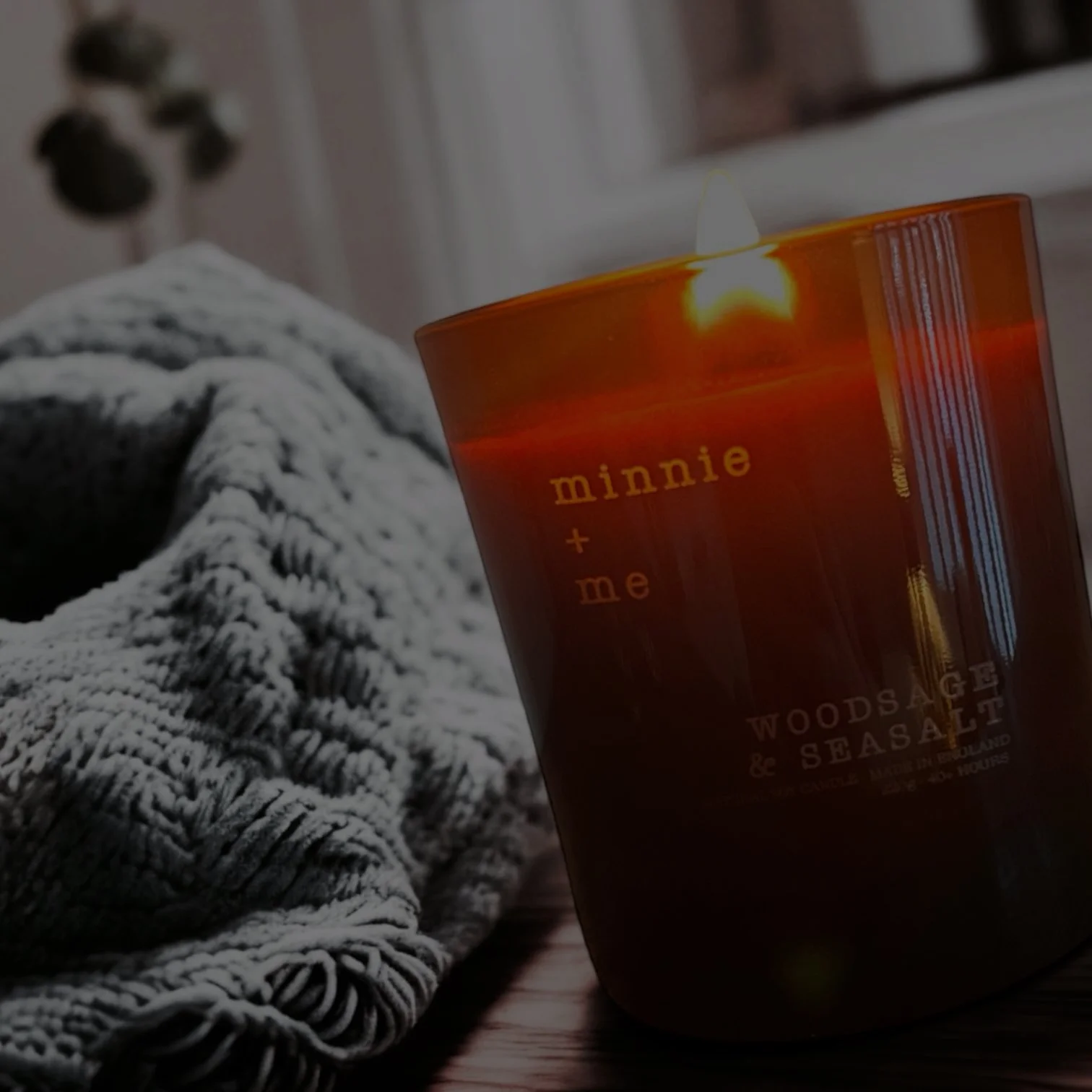 A lit candle in an amber glass jar with the label 'minnie + me' and 'WOODSAW & SEA SALT' on a wooden surface, next to a cozy knitted blanket.