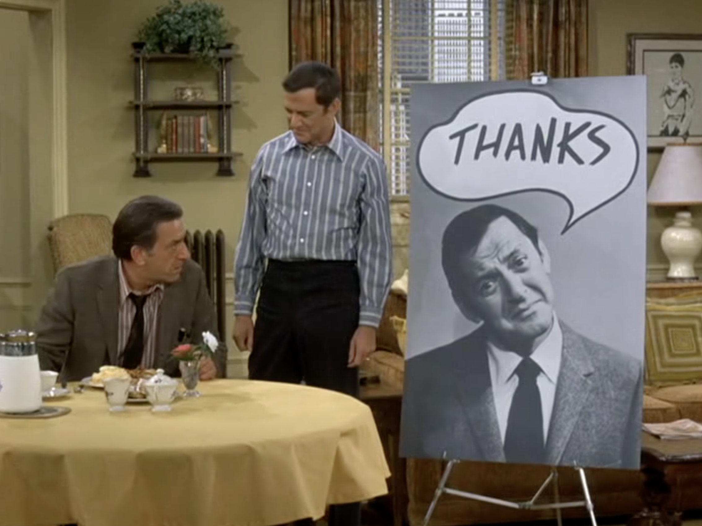 Two men sitting at a dining table with breakfast items, one standing next to a large black and white portrait of a woman with a speech bubble that says 'Thanks'.