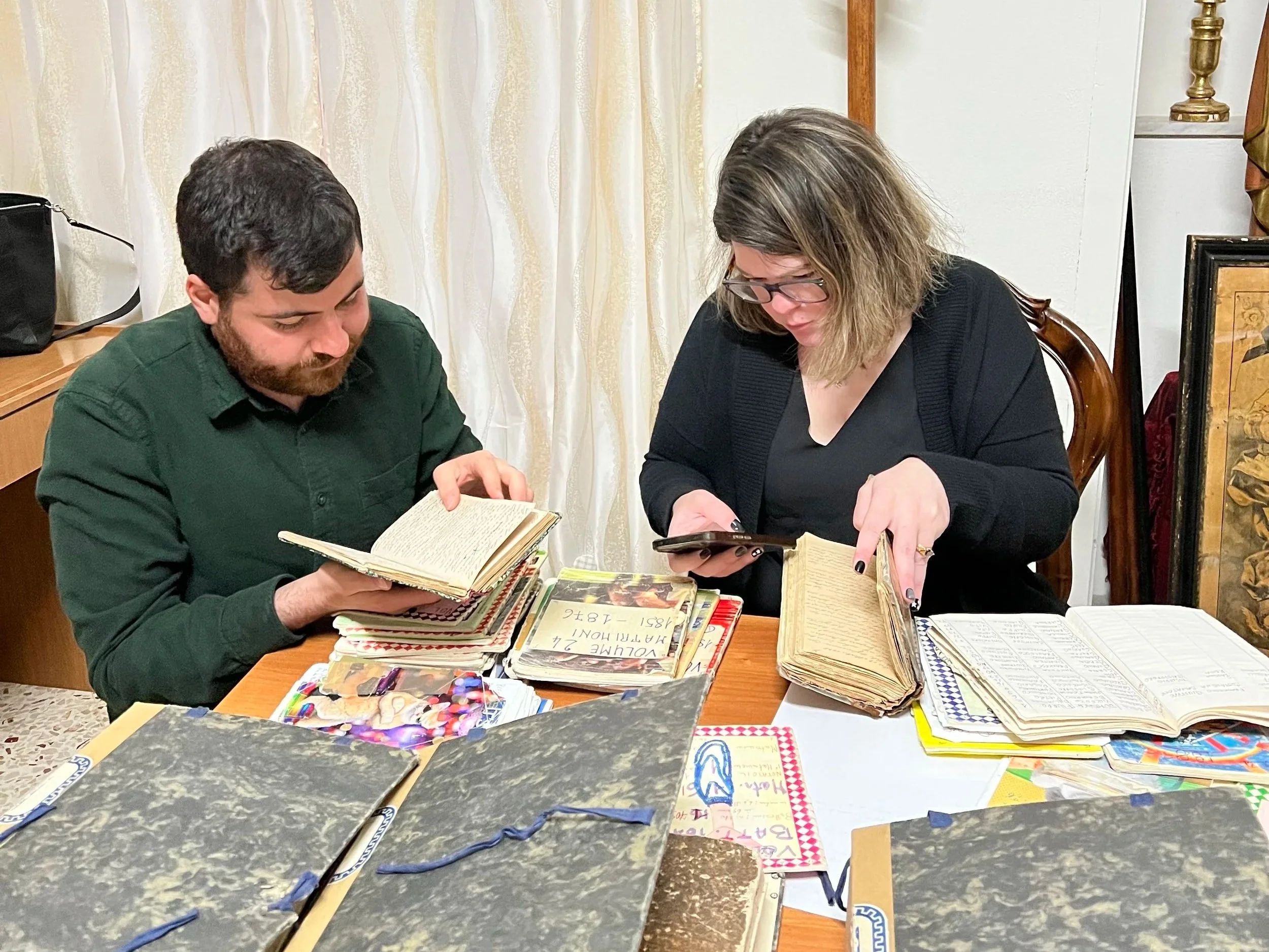 Two people sitting at a table looking at books, with various notebooks and art supplies on the table, in a room with light-colored curtains and decorative items.