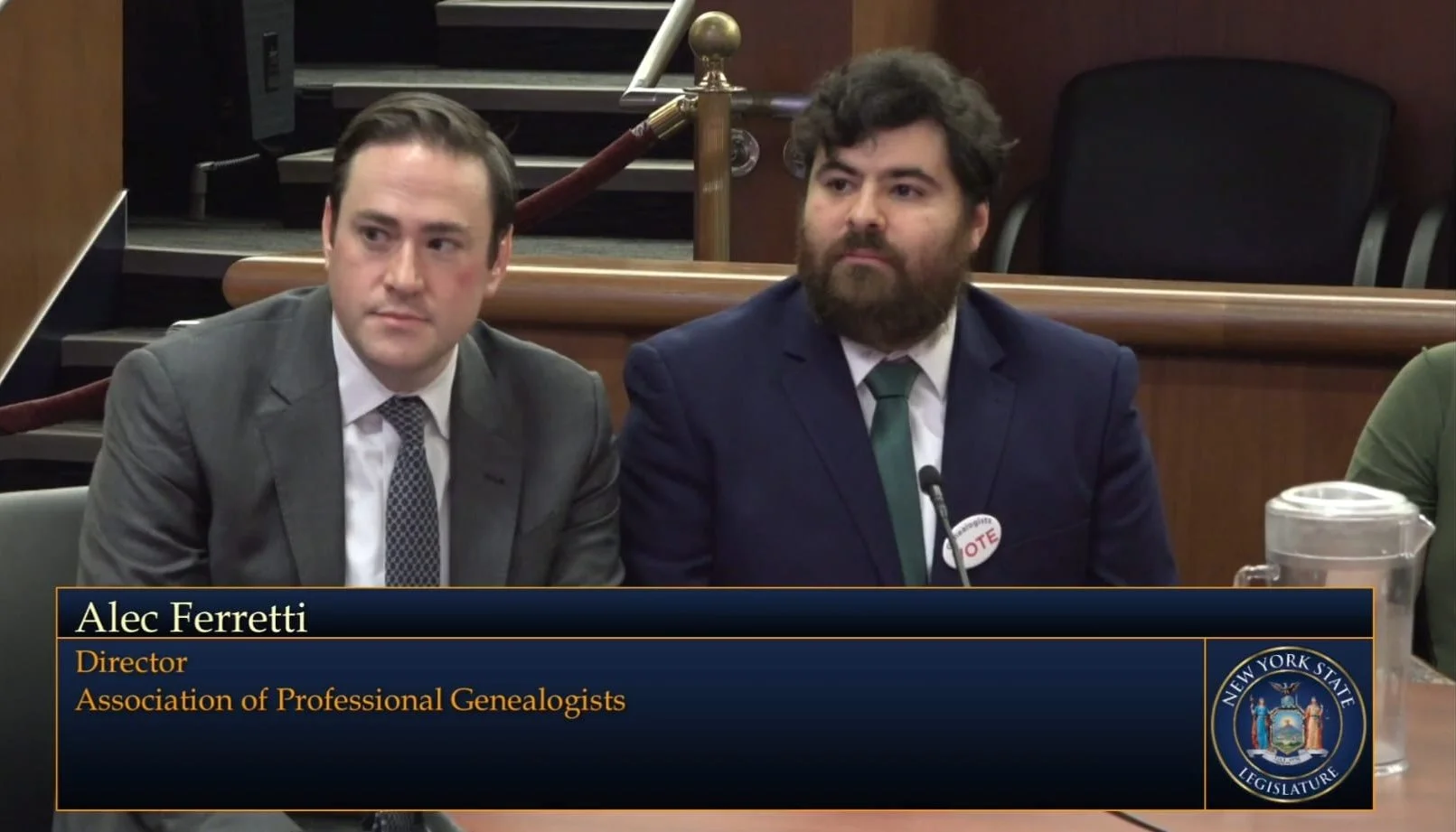 A man in a gray suit and a man in a navy suit sitting at a panel with a sign that reads 'Alec Ferretti, Director, Association of Professional Genealogists,' in a government hearing room with a microphone and a New York State Seal.