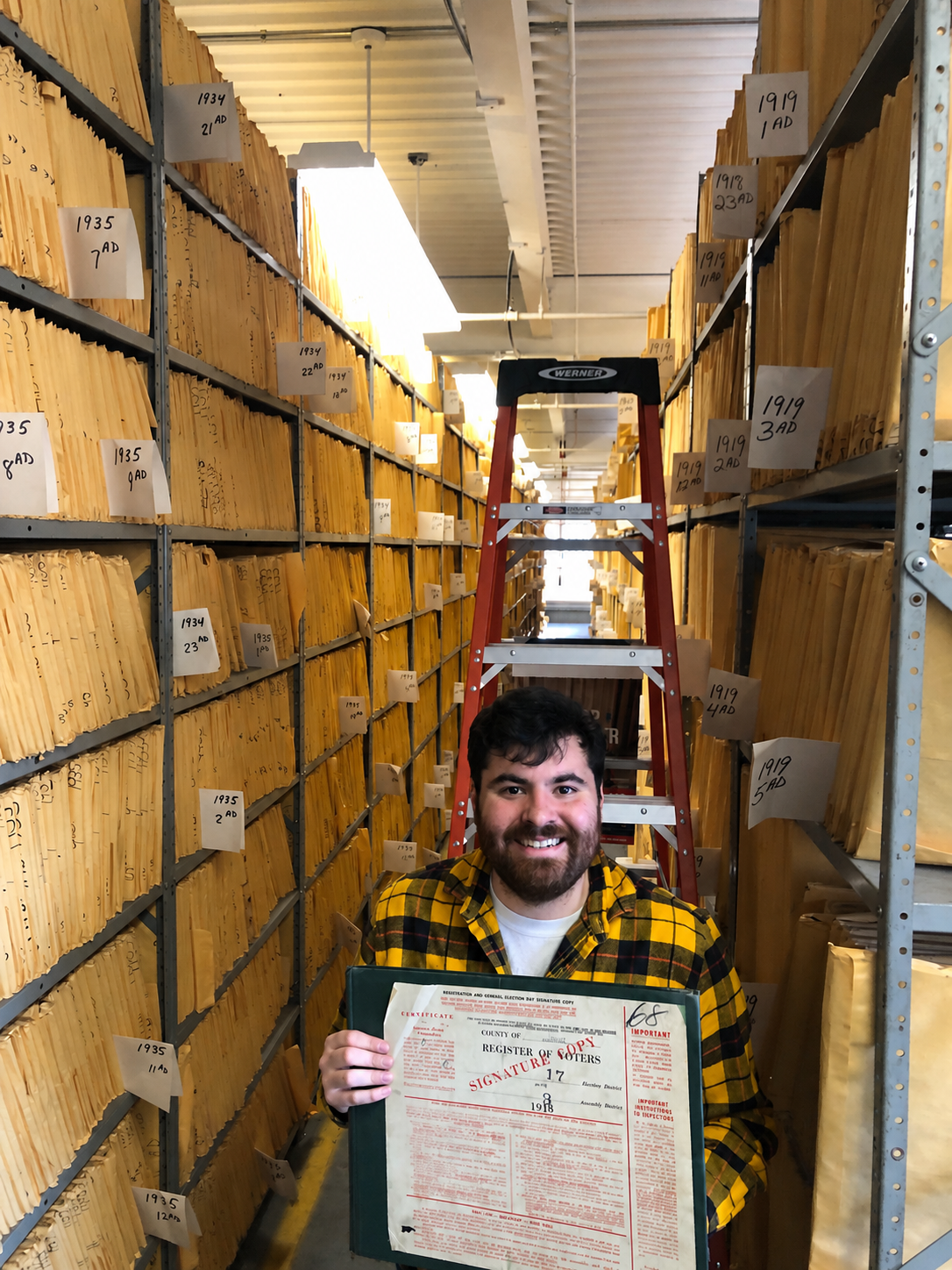 A man smiling, holding a certificate, standing in a storage room with shelves filled with yellow envelopes labeled with years and dates, and a red ladder behind him.