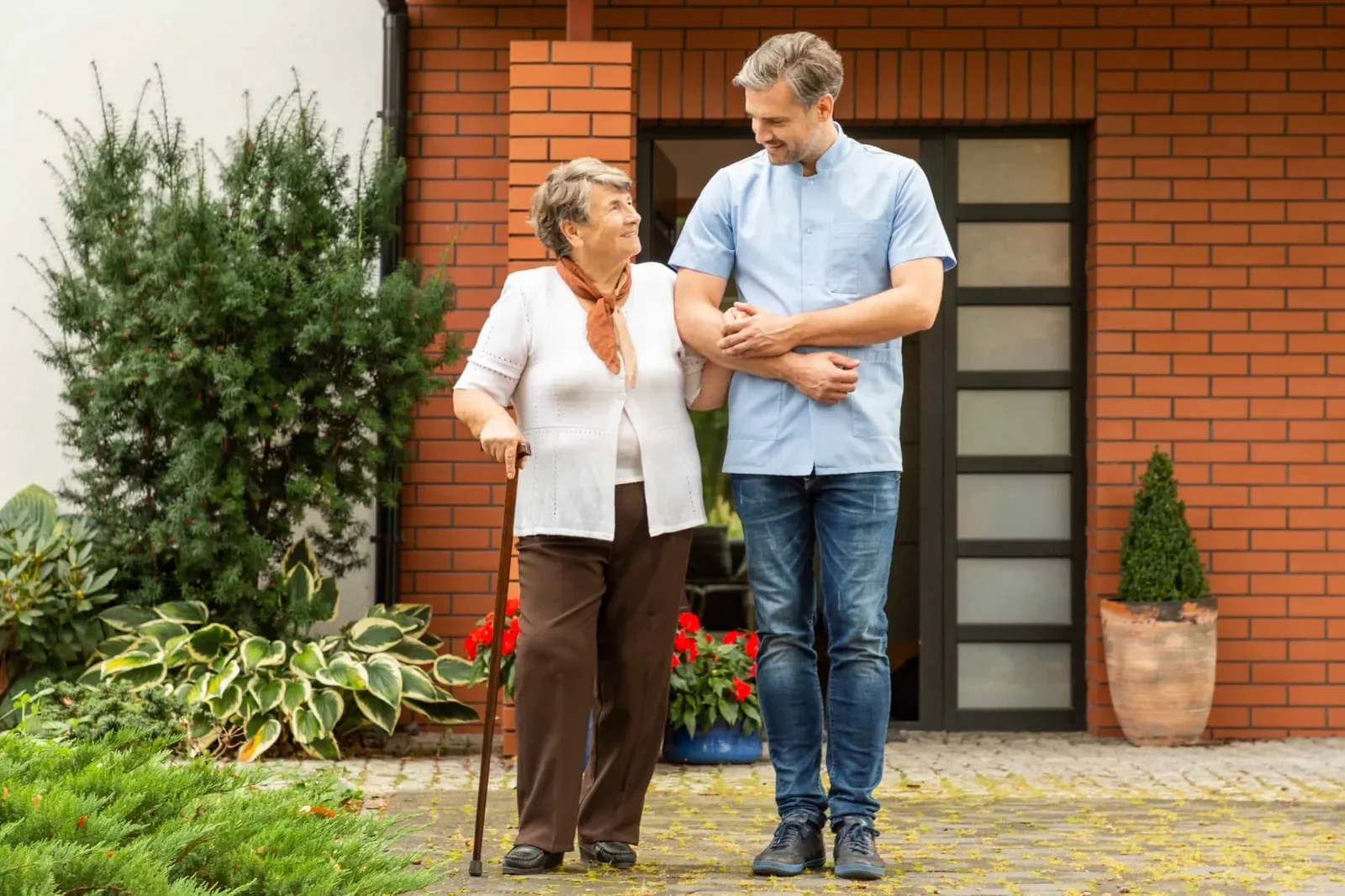 One of Handled Home Care's lovely male caregivers assisting a lady with muscles and its easy for him to help the patient with a bad leg. She is an older adult while walking outside a home in Mesa