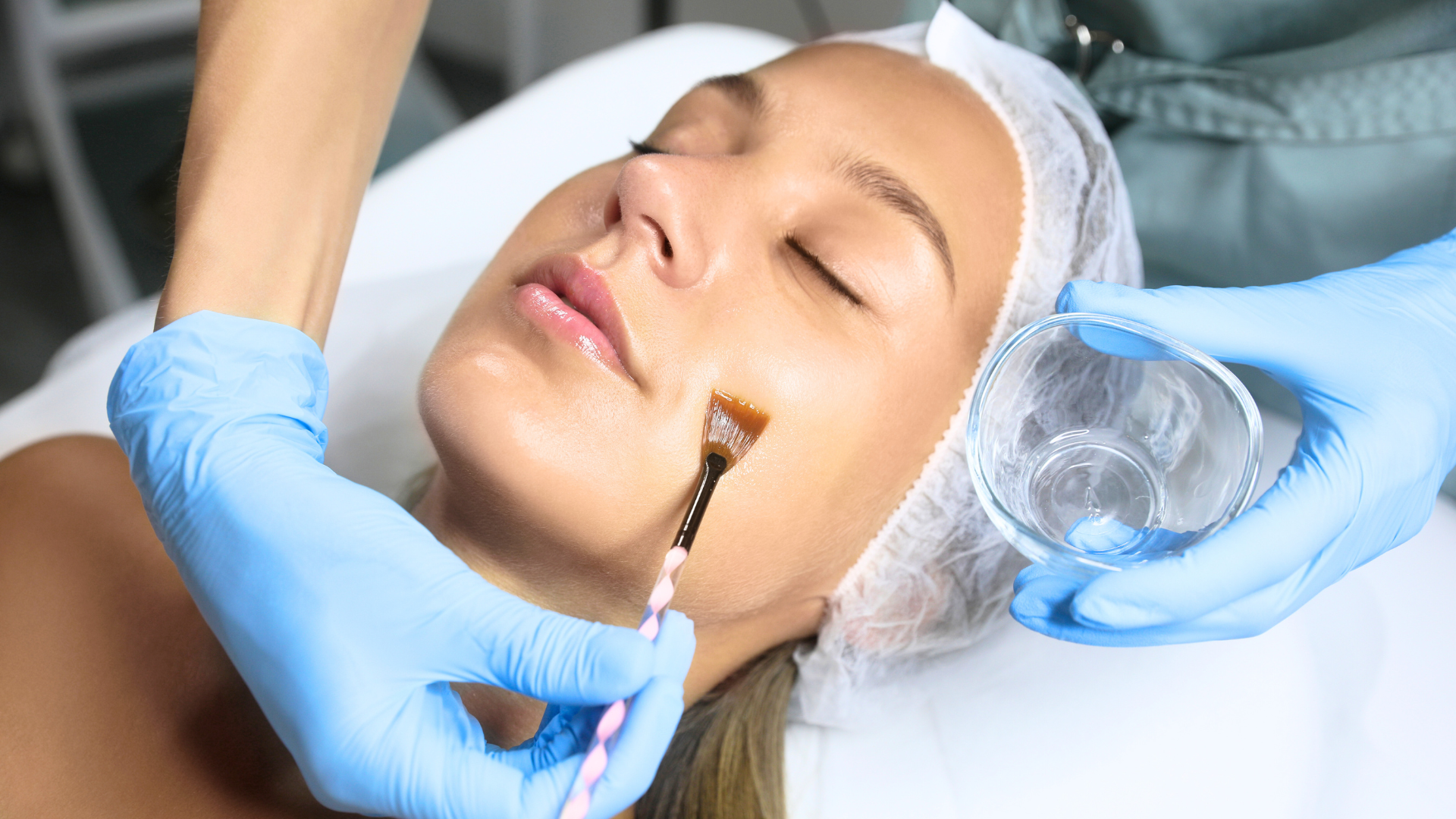 A woman receiving a facial treatment in a spa or skincare clinic, with a professional applying a skincare product on her face with a brush, while holding a bowl of liquid.