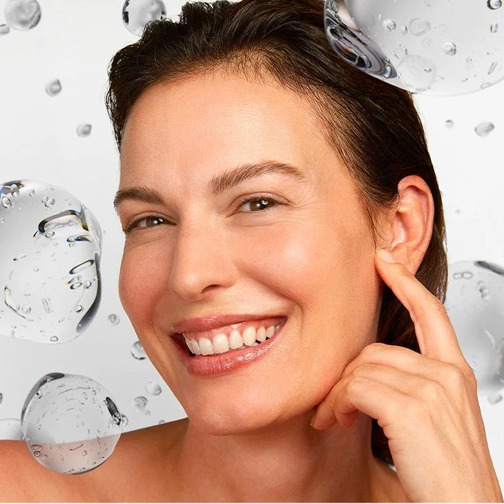 Close-up of a smiling woman with clear skin, touching her ear, with water bubbles floating around her.