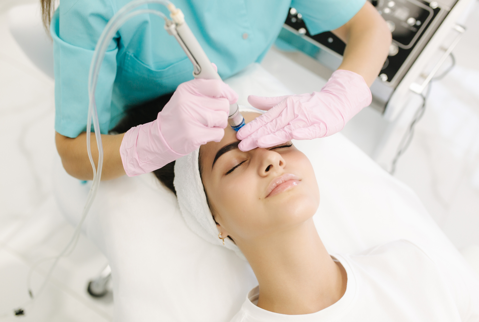 A woman receiving a facial treatment in a clinic, lying on a bed with her eyes closed, while a practitioner with pink gloves uses a device on her forehead.