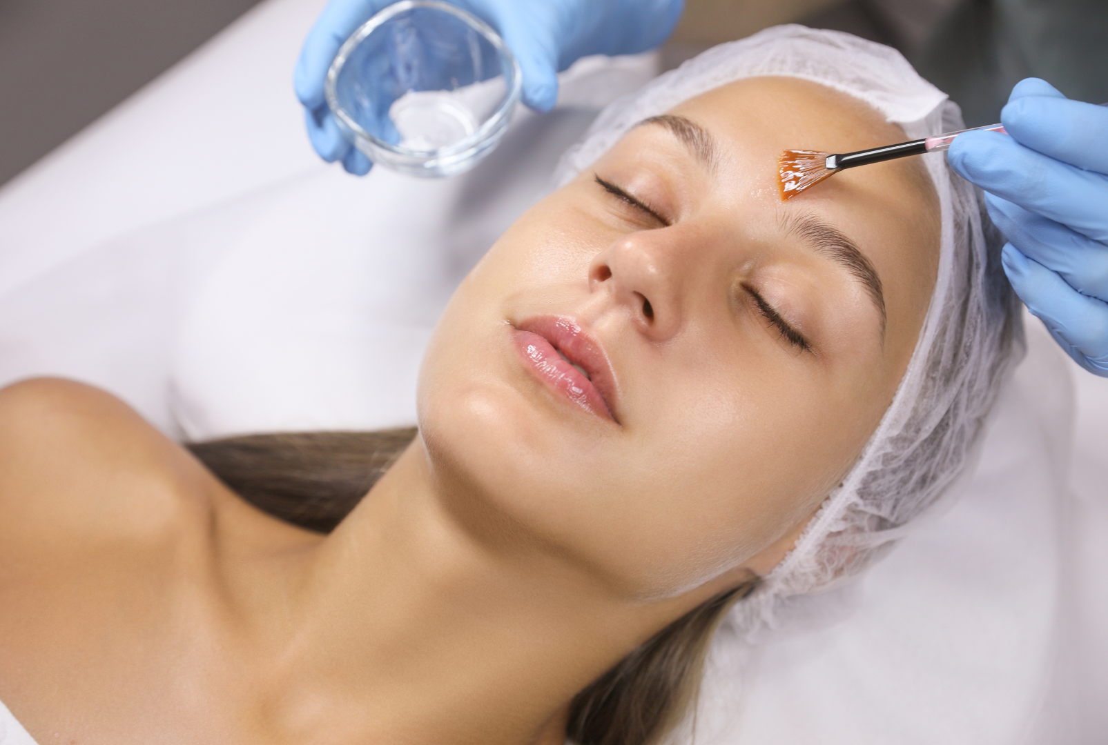 A woman lying down with eyes closed, receiving a facial treatment from a beautician using a brush to apply a substance to her forehead. The woman is wearing a hair cap and has a calm expression.