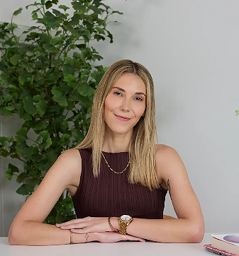 Young woman with blonde hair wearing a sleeveless maroon top and gold watch, sitting at a white desk with a plant in the background.